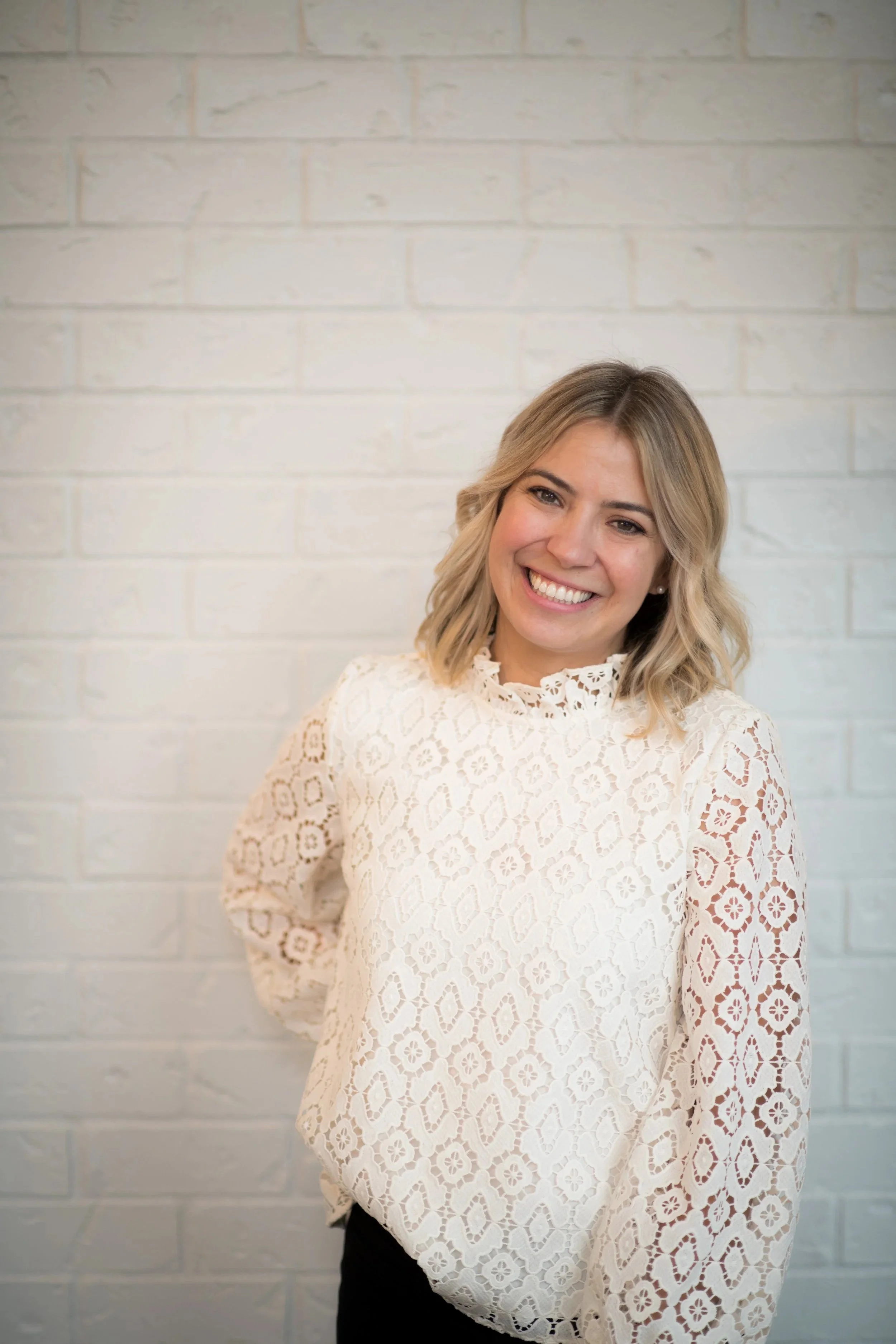 A woman smiling, with blonde, wavy hair, wearing a white lace long-sleeved top with floral patterns, standing against a light brick wall.