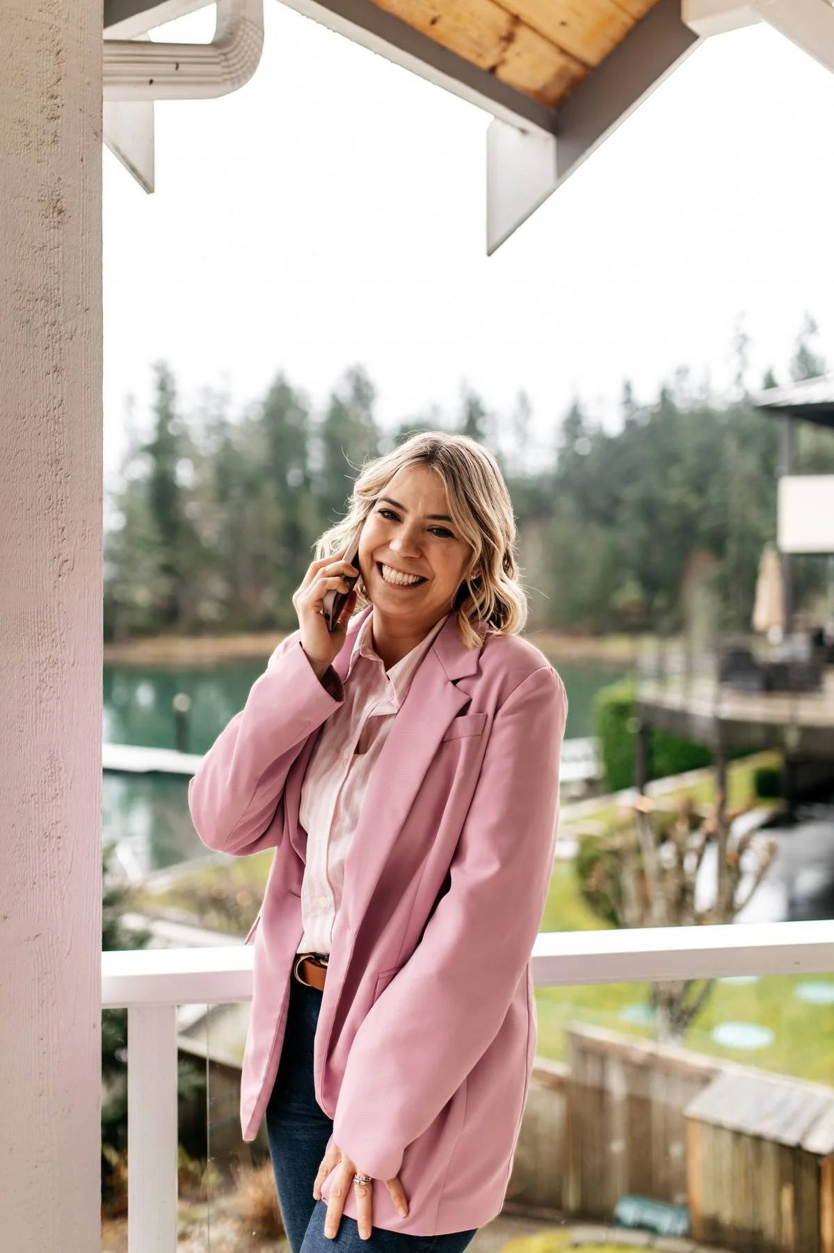 A woman in a pink blazer and white shirt talking on a cell phone, standing on a porch outdoors with trees and water in the background.