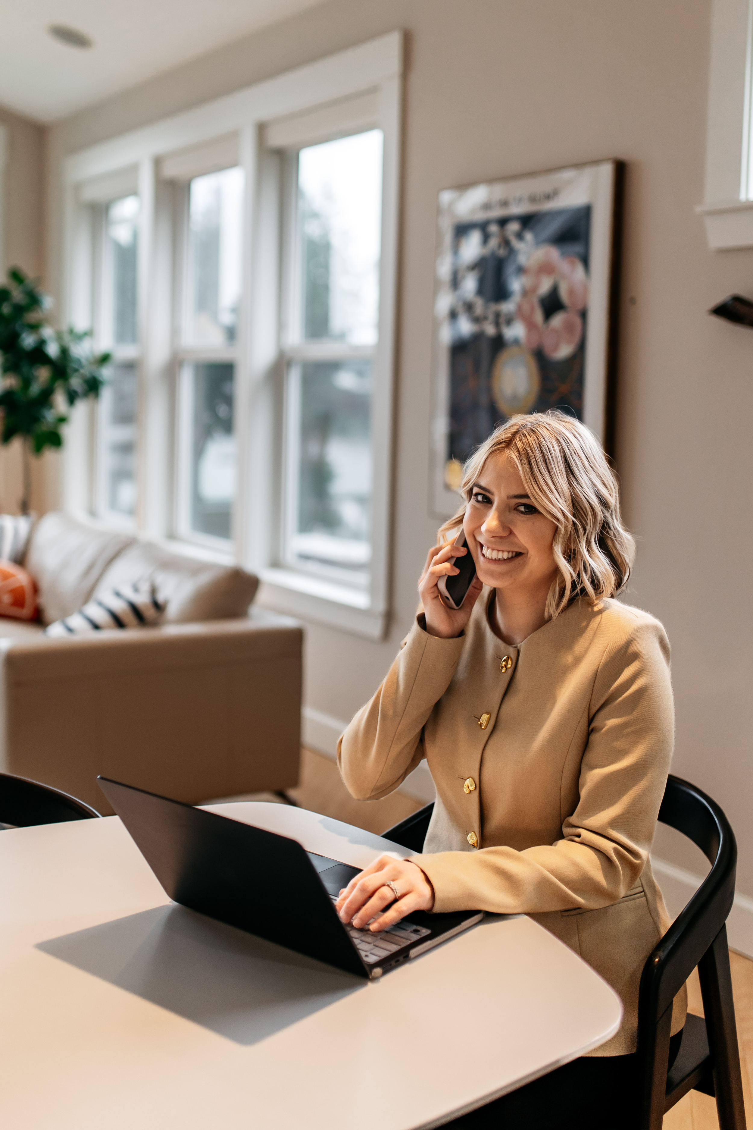 Woman in beige blazer smiling and talking on a landline phone while sitting at a desk with a laptop in a bright living room with large windows, a beige sofa, and framed artwork on the wall.