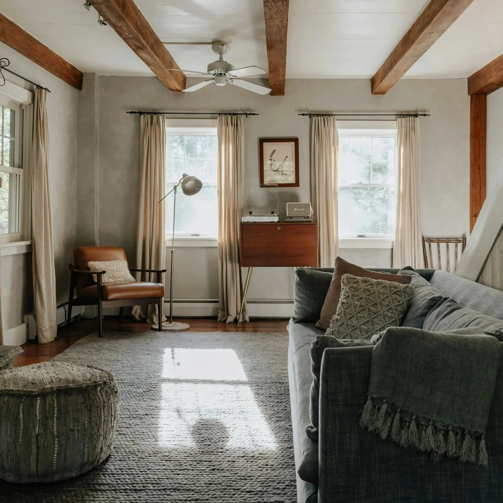 Living room with beige curtains on windows, gray sofa with textured pillows, a vintage wooden sideboard with a stereo and speaker, a leather armchair with a throw pillow, a floor lamp, wooden ceiling beams, and a textured area rug.