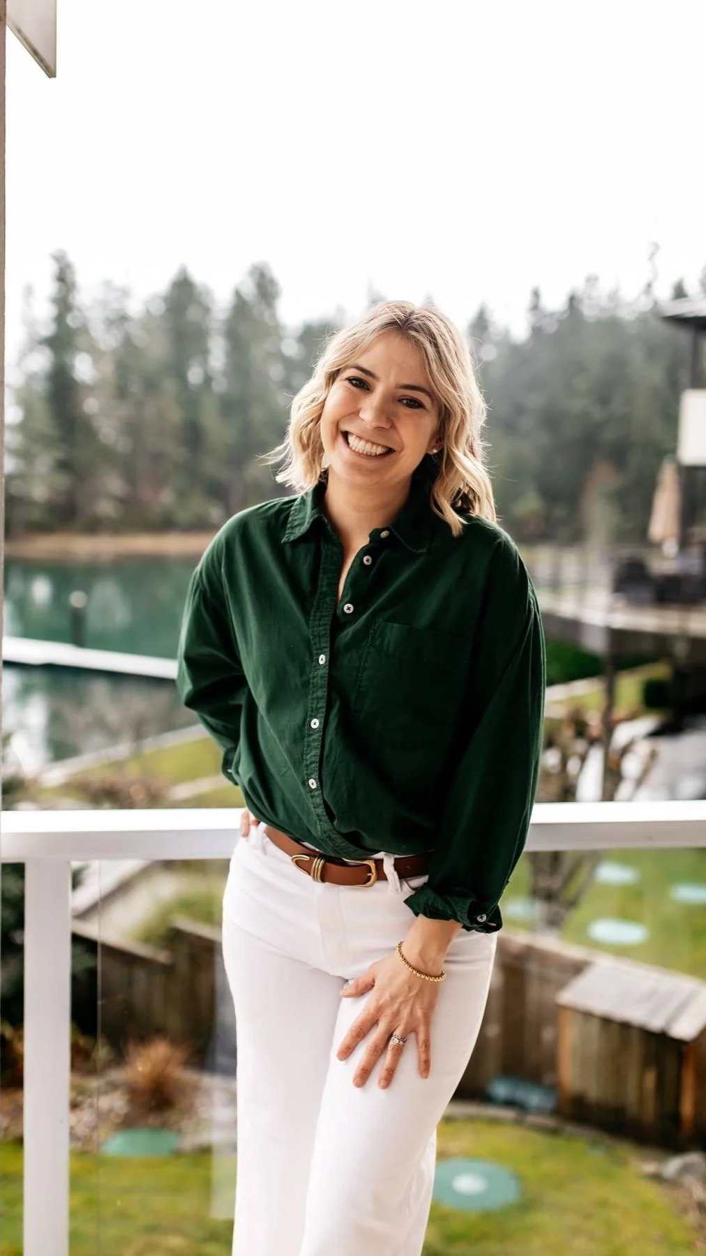 Smiling young woman with blonde hair, green shirt, and white pants standing outdoors near water with trees in the background.