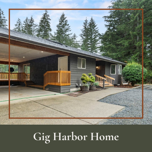 A single-story house with gray siding, a small porch with wooden railings, surrounded by trees and plants, with a driveway and gravel area in front.