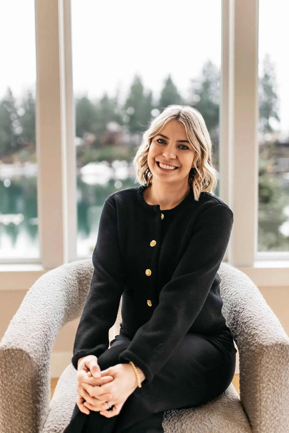 A young woman with blonde, shoulder-length wavy hair, smiling, sitting on a plush beige armchair with a textured fabric. She is wearing a black cardigan with gold buttons, black pants, and a bracelet on her left wrist. She is in a bright room with large windows and a view of trees and water outside.