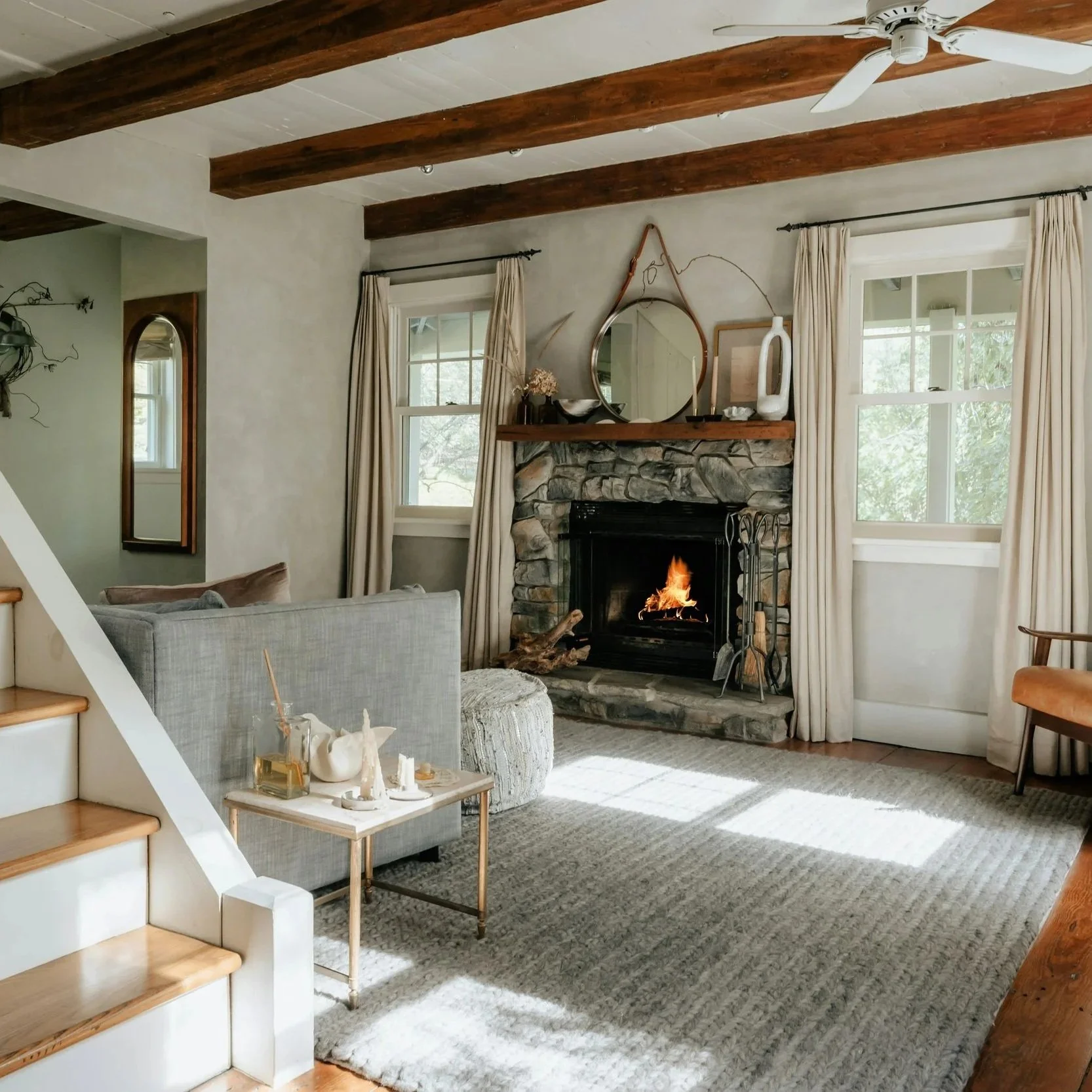 Living room with stone fireplace, wooden beams on ceiling, two windows with cream curtains, sofa, ottoman, and coffee table with decorative items, ceiling fan, and beige carpet.