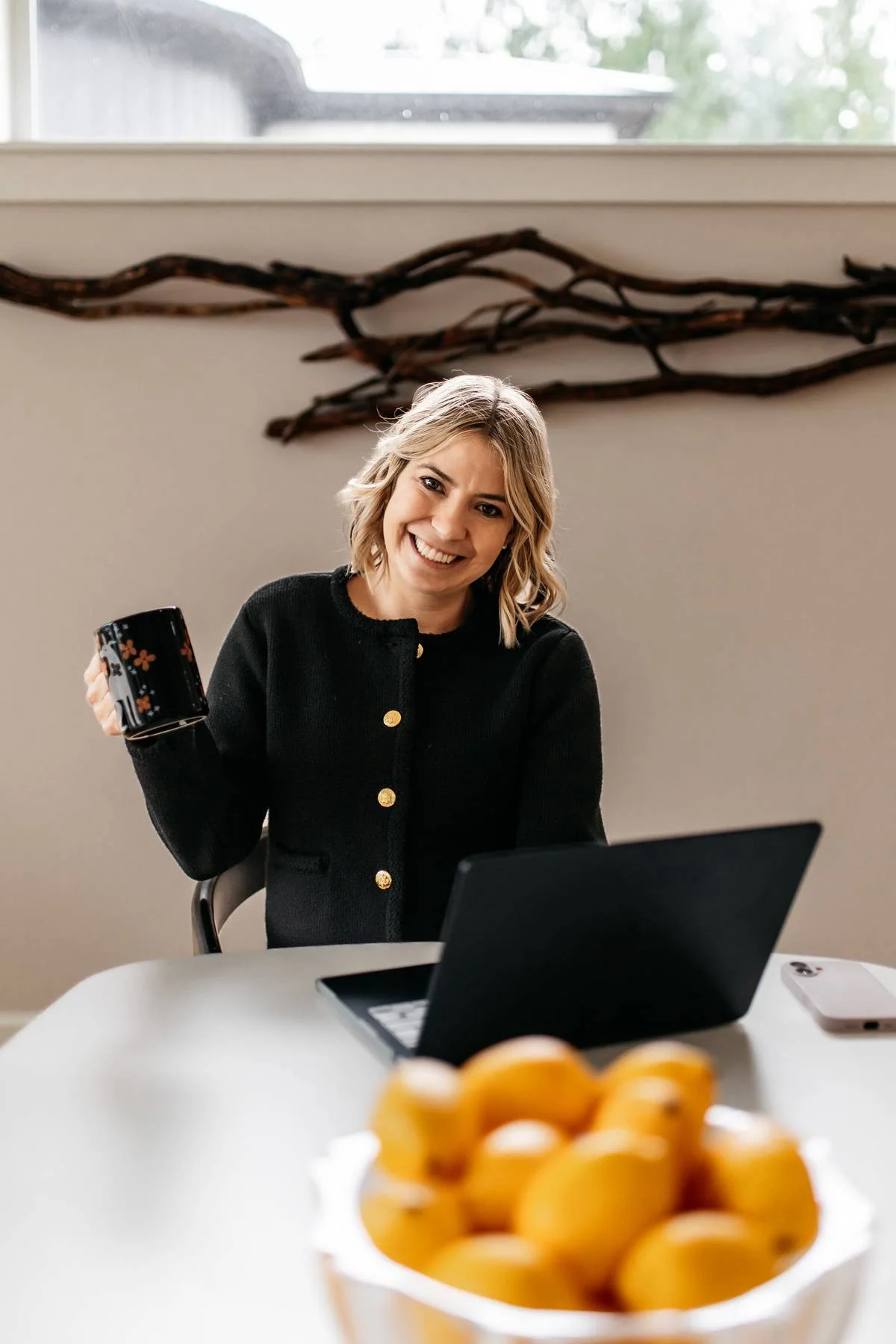 Woman with blonde hair smiling, holding a black coffee mug, sitting at a table with a laptop, in a bright room with a window, and a bowl of apricots in the foreground.