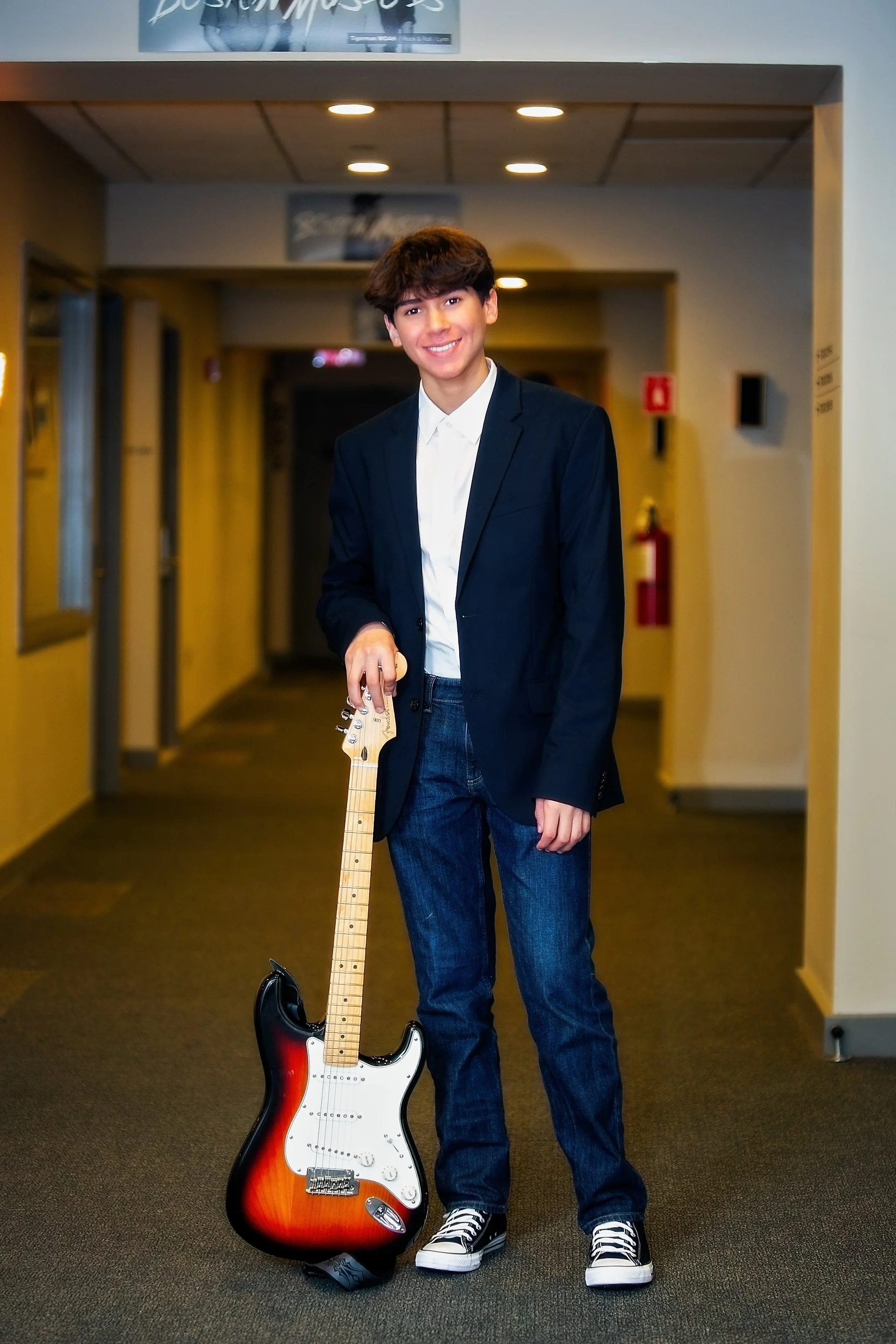 Young man in a navy blazer and jeans standing in an indoor hallway holding a sunburst electric guitar.