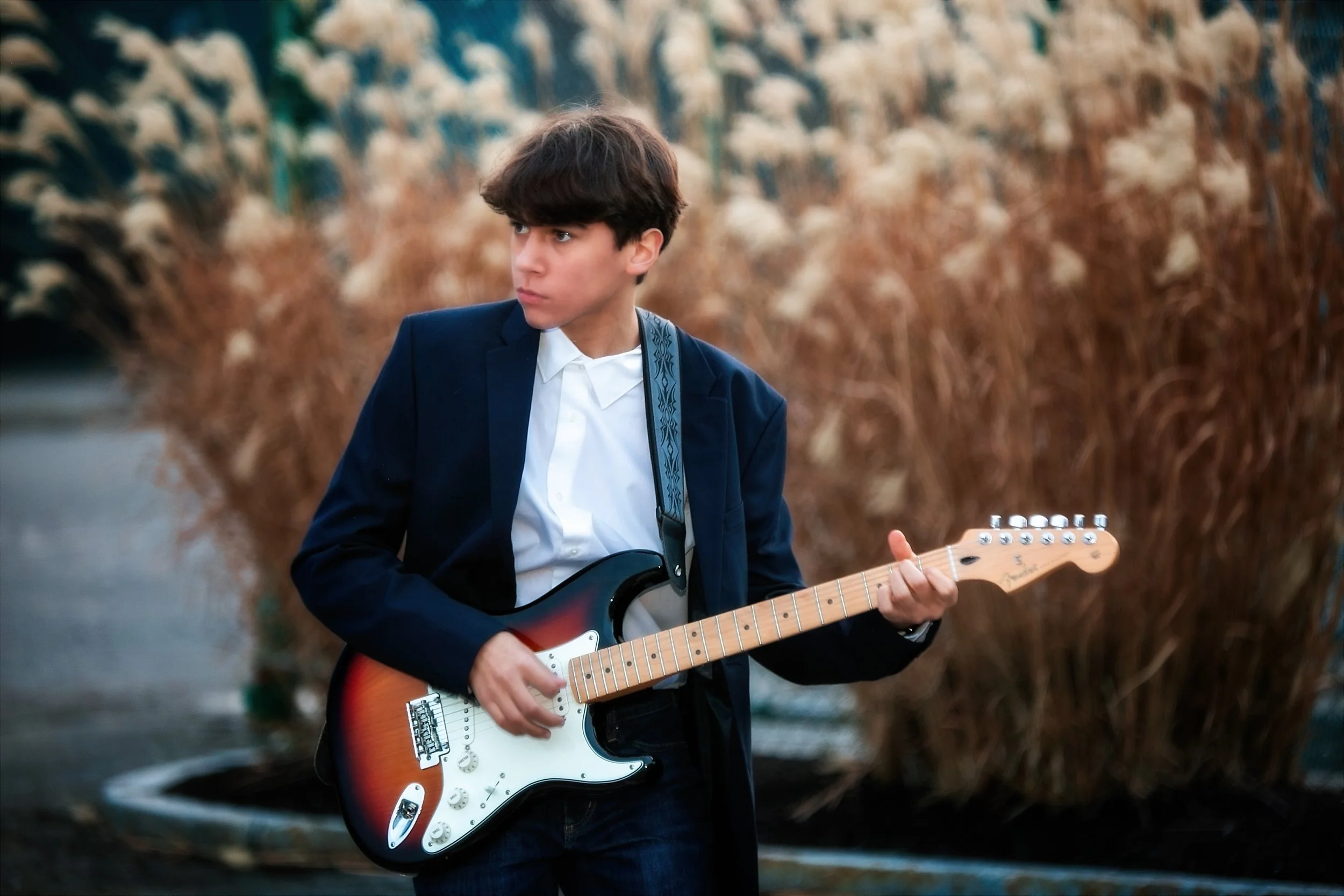 A young man with dark hair wearing a dark blazer and white shirt playing an electric guitar outdoors, with tall dried plants or weeds in the background.