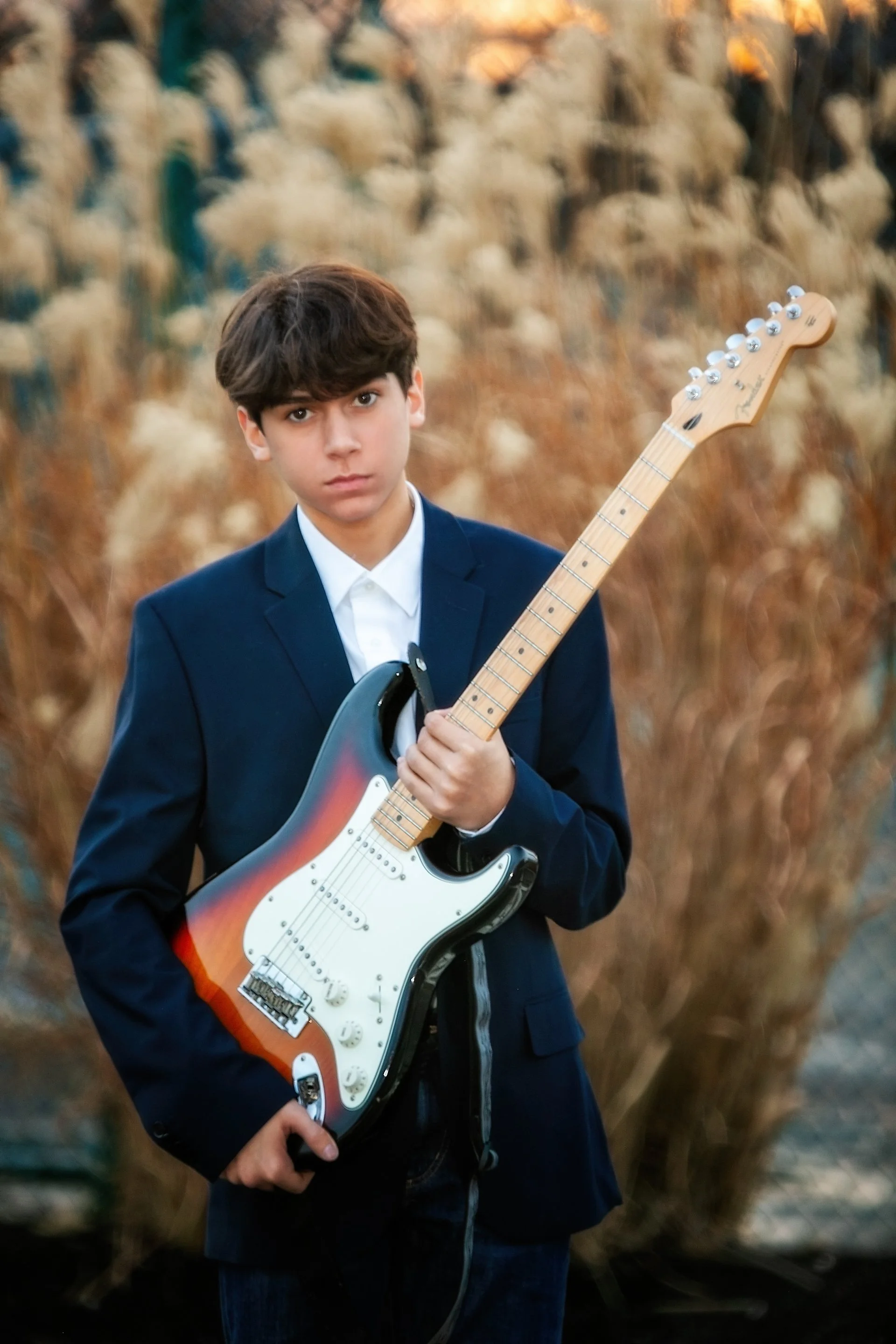 Young boy in a blue blazer holding an electric guitar outdoors, with tall dry grass in the background during sunset.