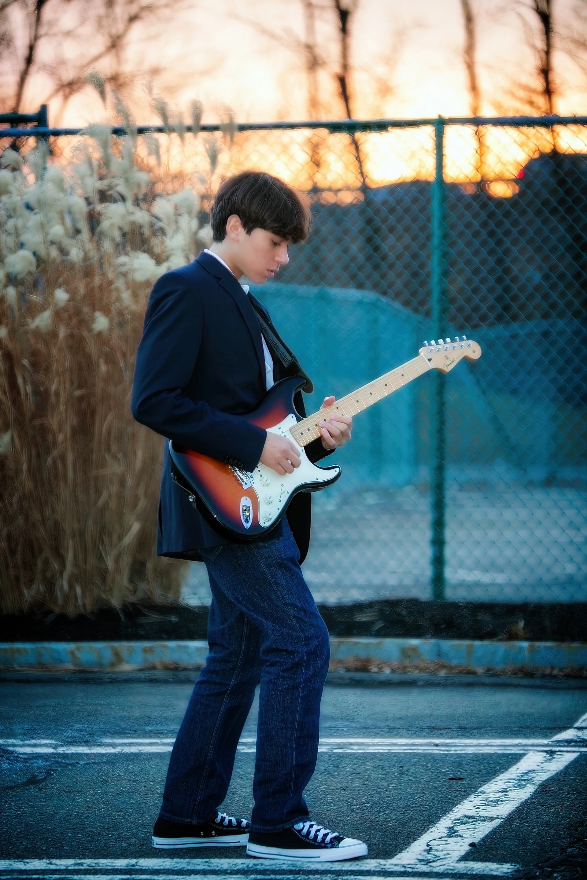 A young person with dark hair wearing a blue blazer, jeans, and black sneakers playing an electric guitar in a parking lot at sunset. The background includes a chain-link fence and trees with a colorful sky.