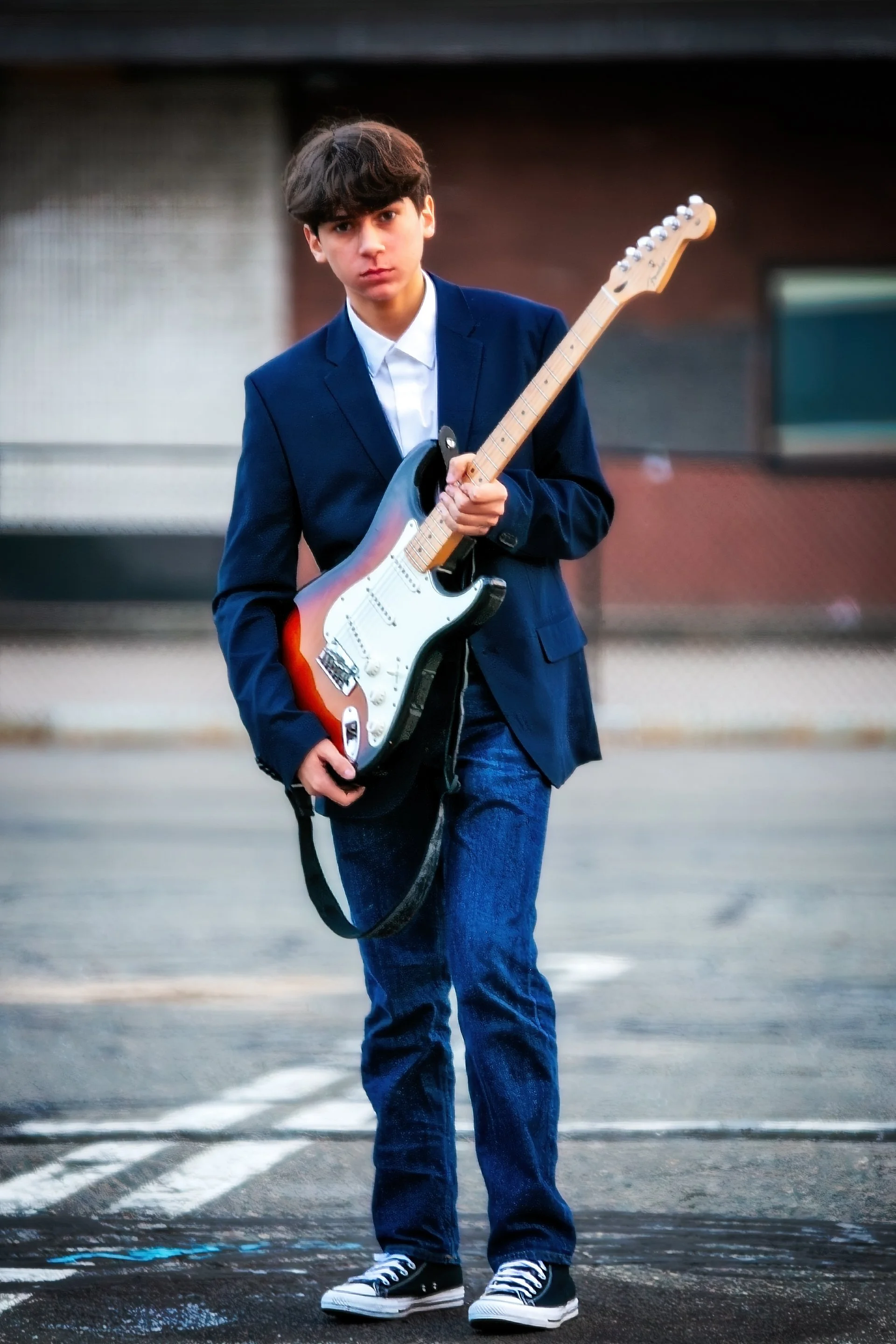 Young man in a blue suit and jeans holding an electric guitar standing on an empty street.