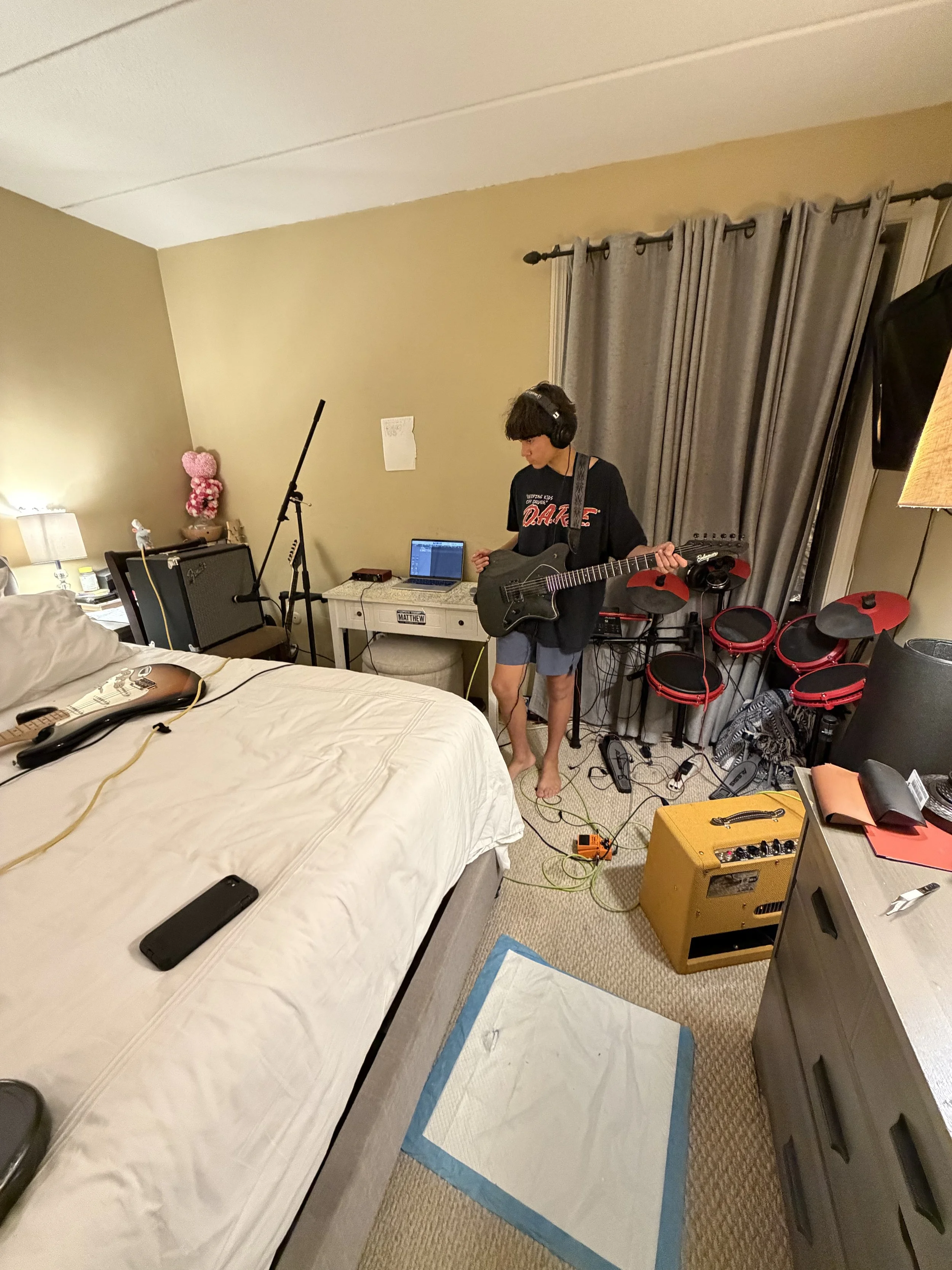 A person playing an electric guitar in a cluttered bedroom with a bed, electronic equipment, and musical instruments, including a yellow amplifier, a drum set, and a laptop on a nightstand.