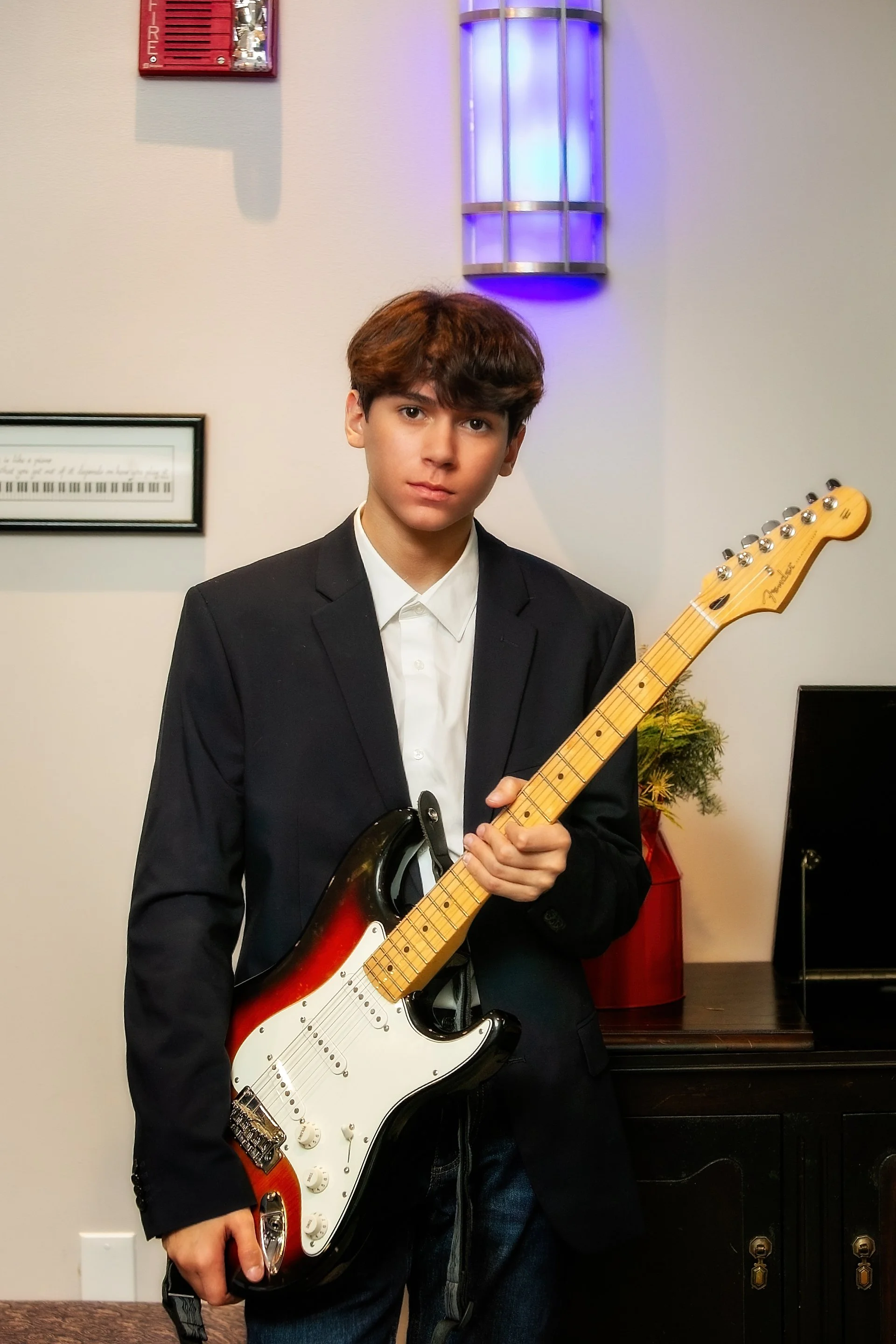 Young boy in a black blazer holding a Fender Stratocaster electric guitar, standing indoors with a wall-mounted fire alarm and purple wall sconce in the background.
