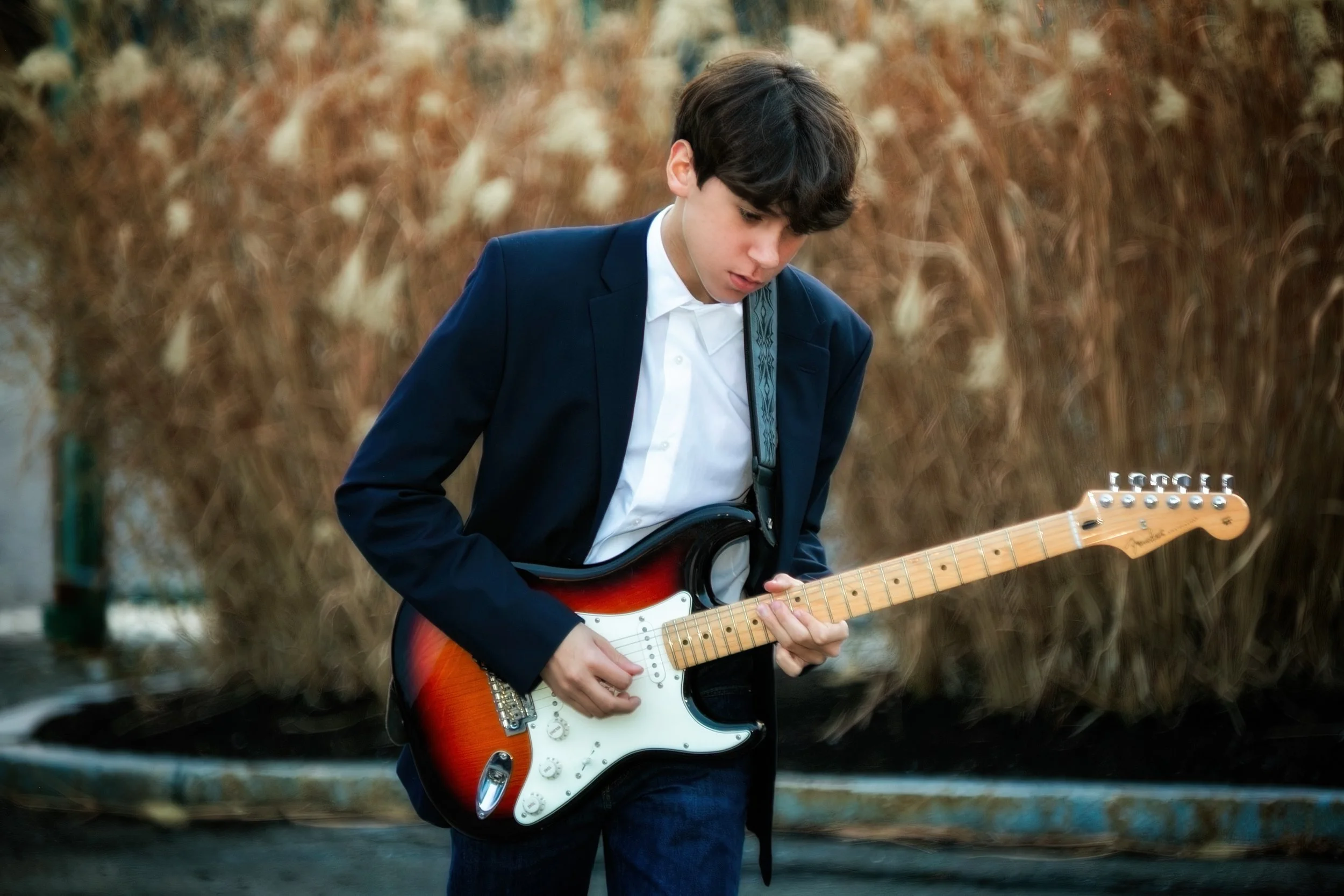 A young boy with dark hair wearing a blue blazer and white shirt playing an electric guitar outdoors with brown and beige foliage in the background.