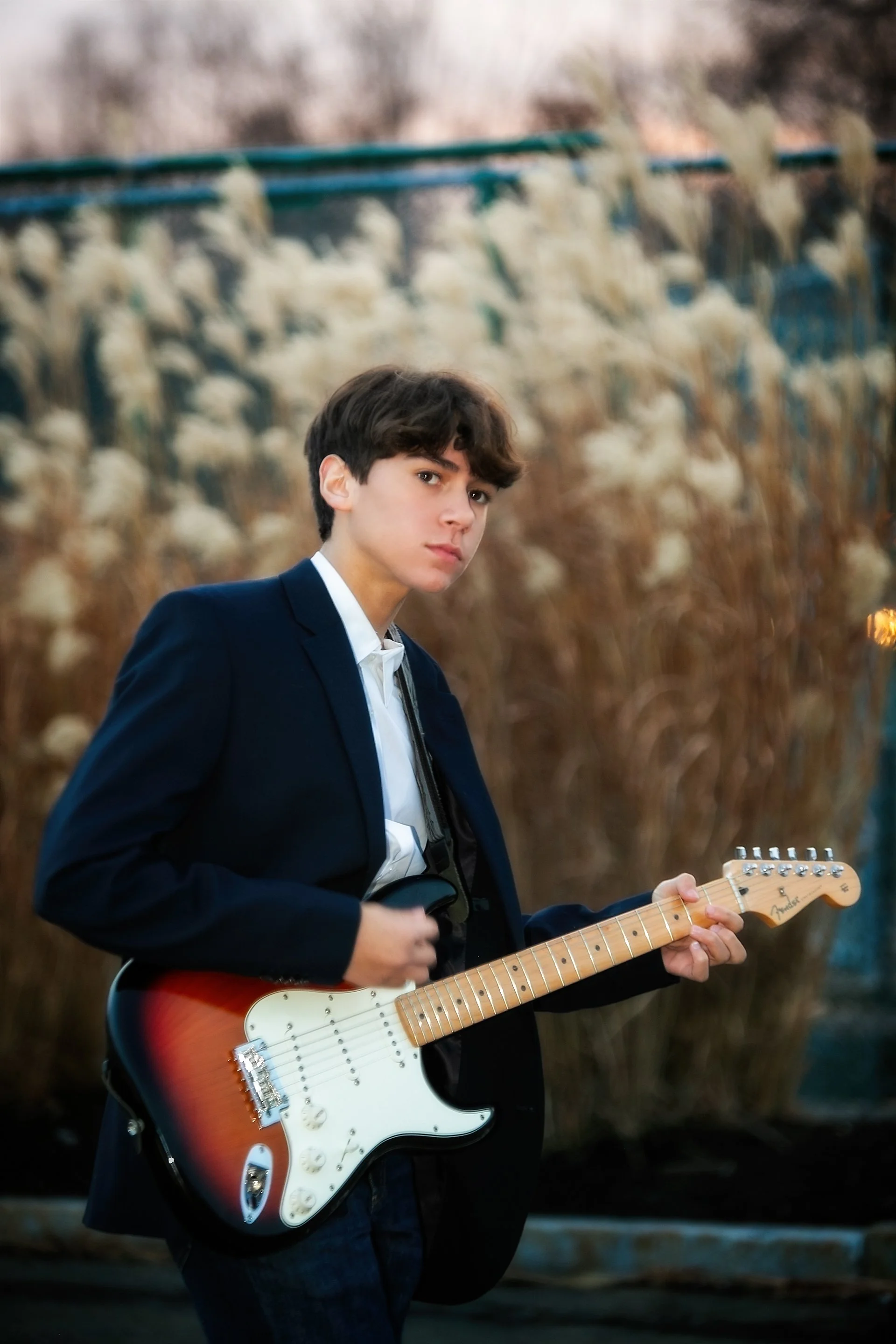 A young man in a black blazer and white shirt holding an electric guitar outdoors with a blurred background of plants and trees.