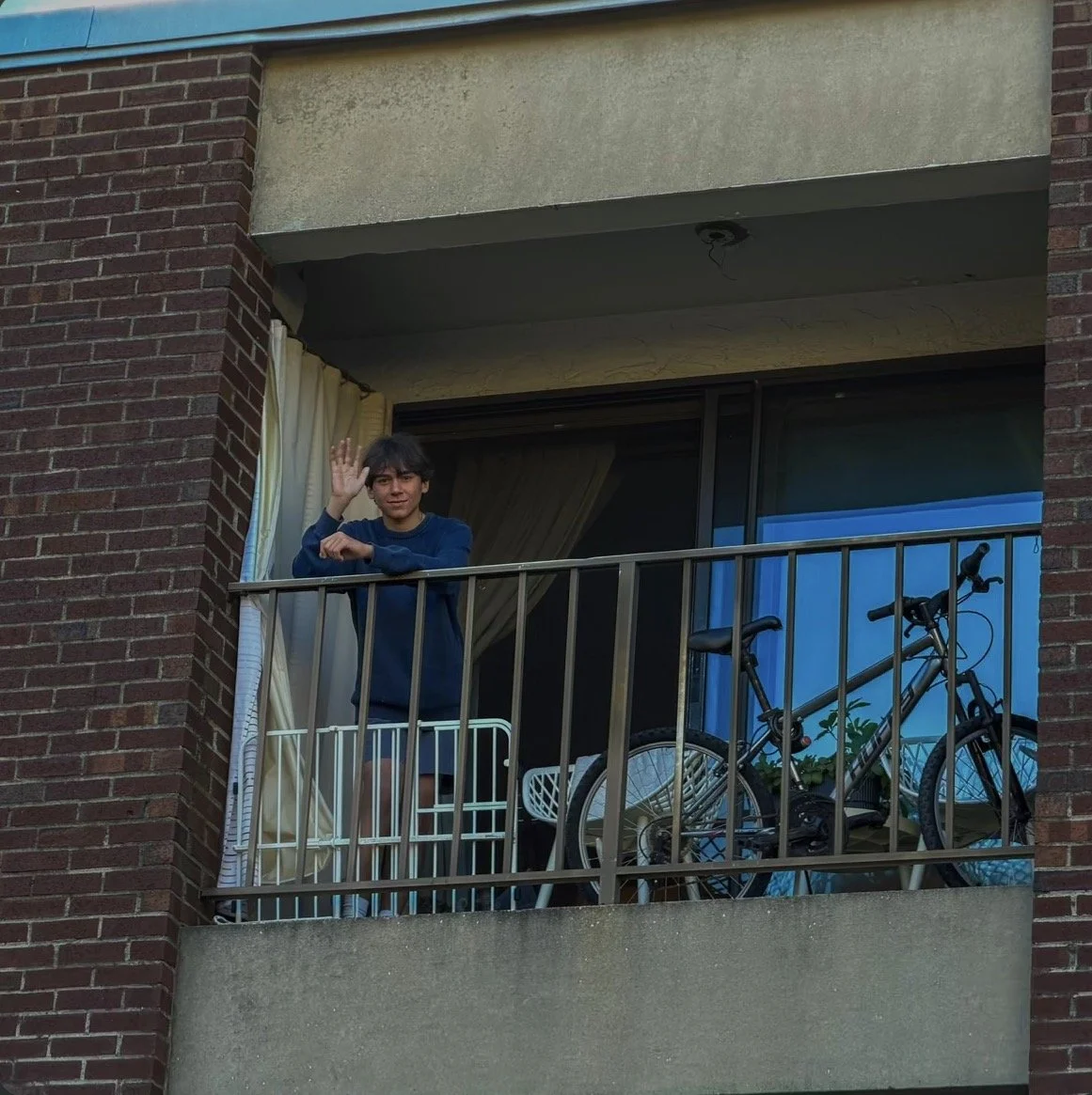 A young person standing on a balcony, waving, with bicycle, laundry rack, and plants behind them.