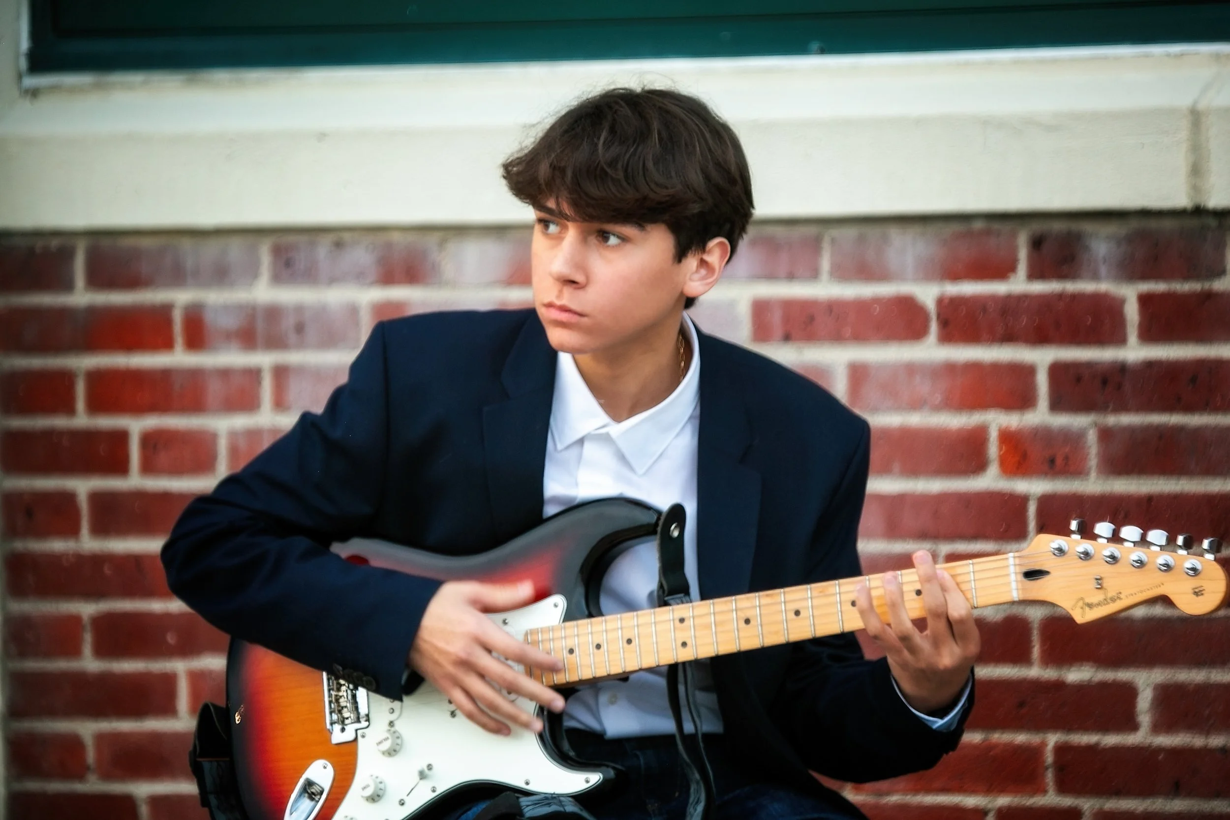 Young man wearing a dark blazer and white shirt playing an electric guitar in front of a brick wall.
