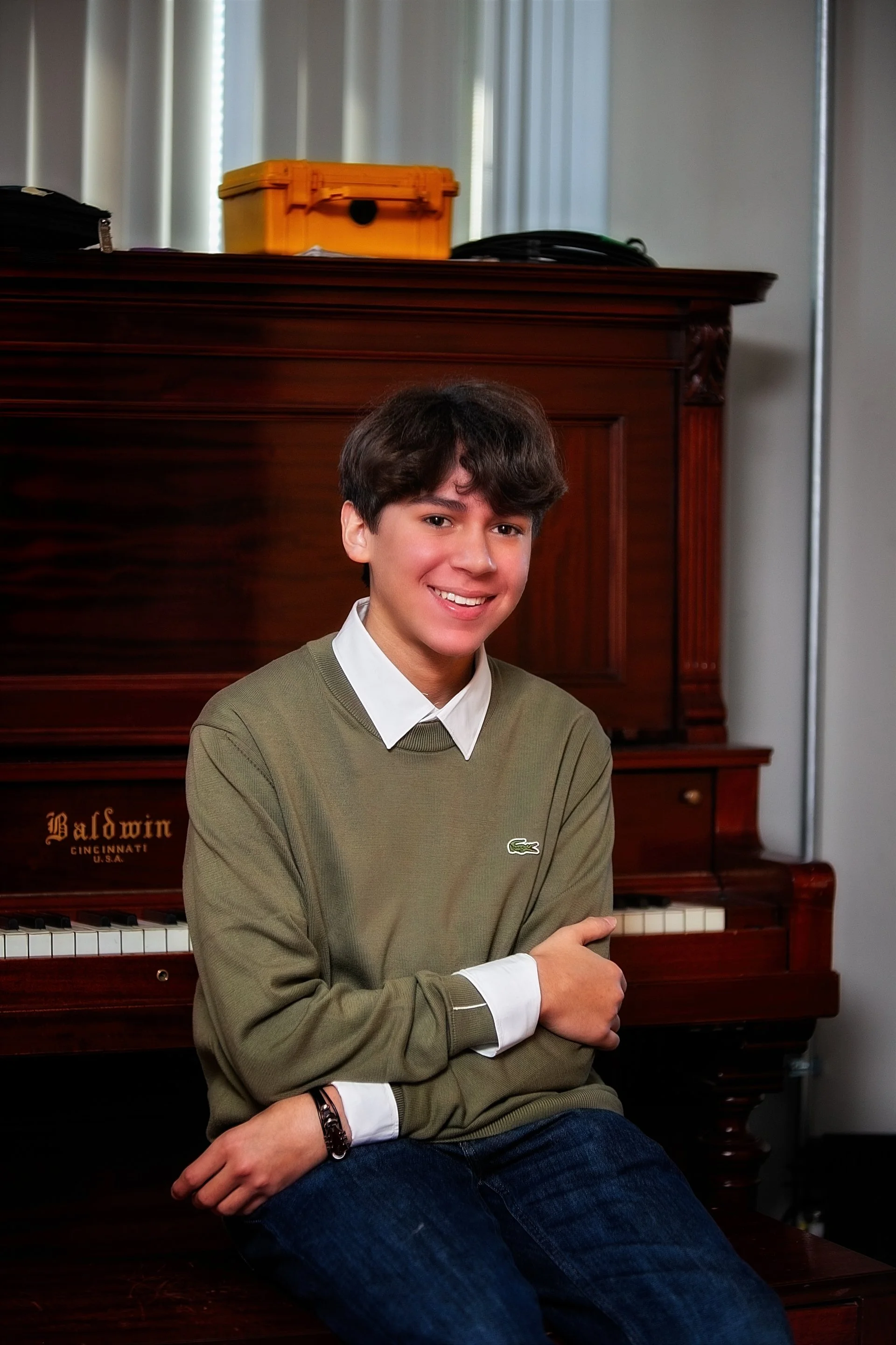 A young man with brown hair, wearing a green sweater and white collared shirt, sitting in front of a Baldwin upright piano, smiling at the camera.