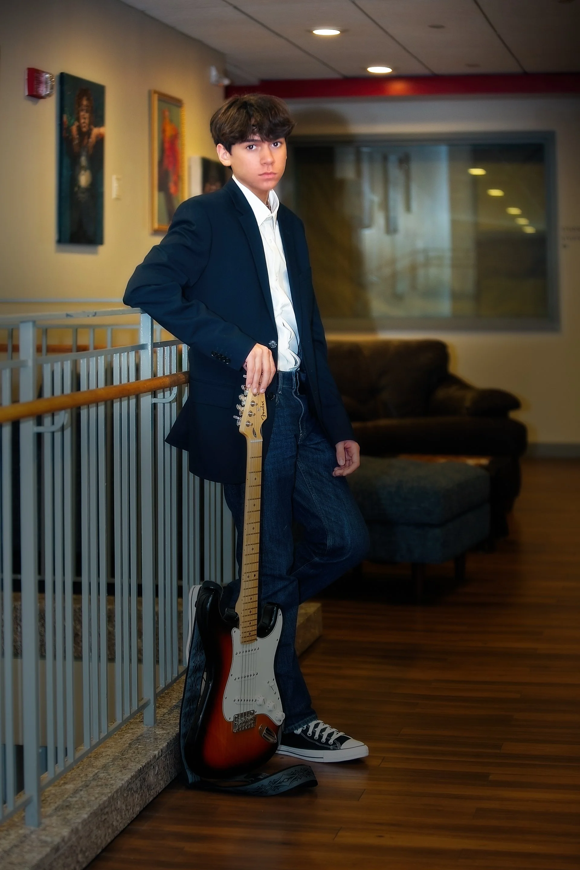 Young boy standing indoors, leaning on a railing, holding an electric guitar, wearing a dark blazer, white shirt, jeans, and sneakers, with paintings on the wall and a large window in the background.