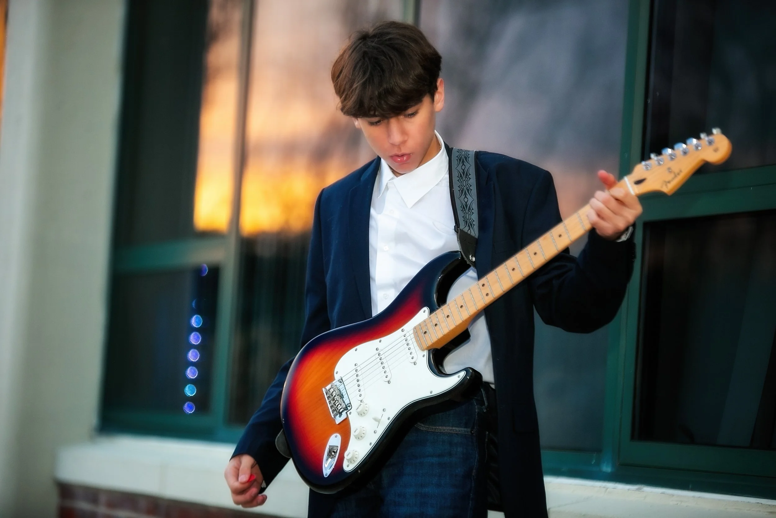 A young boy with brown hair wearing a white shirt and dark jacket playing an electric guitar in an indoor space with a reflective wall.