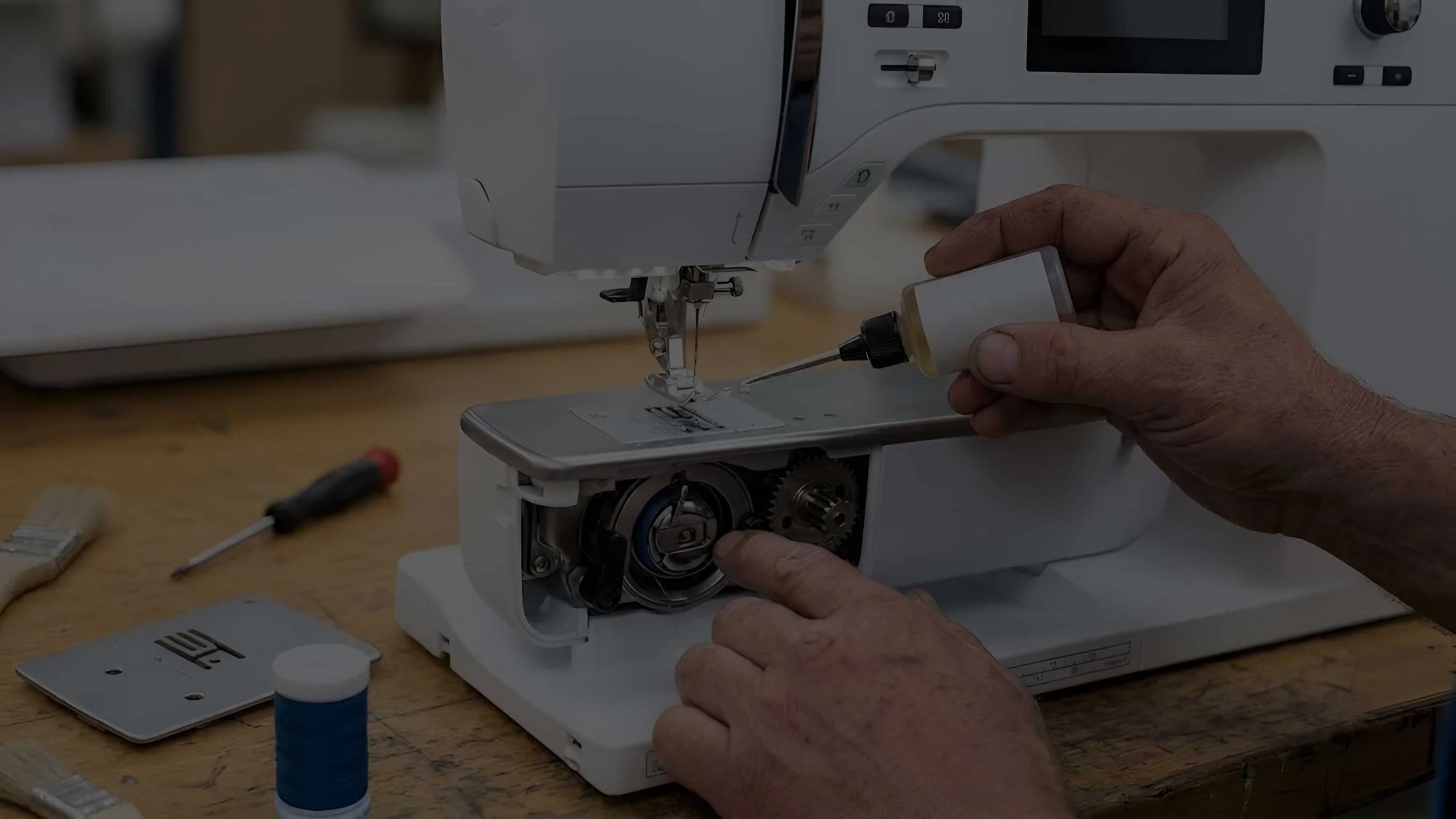A person repairing a sewing machine, holding a tool and working on the machine's internal components, with various tools and sewing materials on the table.