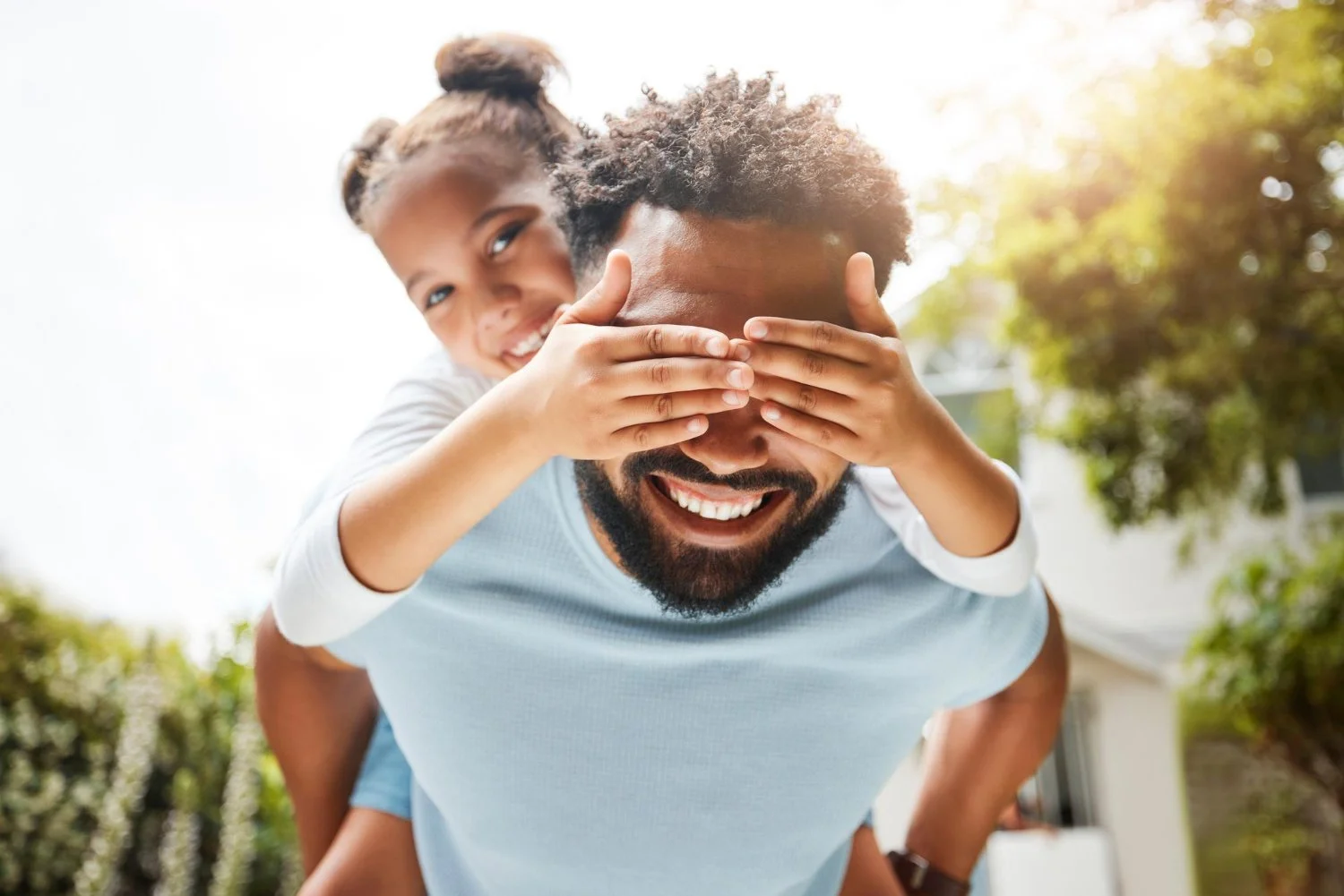A man giving a piggyback ride to a young girl outside on a sunny day, with the girl smiling and covering the man's eyes with her hands.