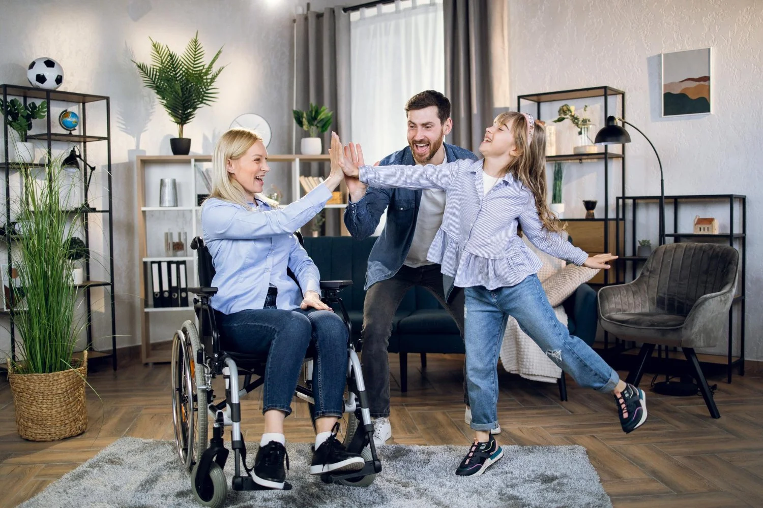 Family celebrating in living room: woman in wheelchair clapping with girl, man smiling between them.