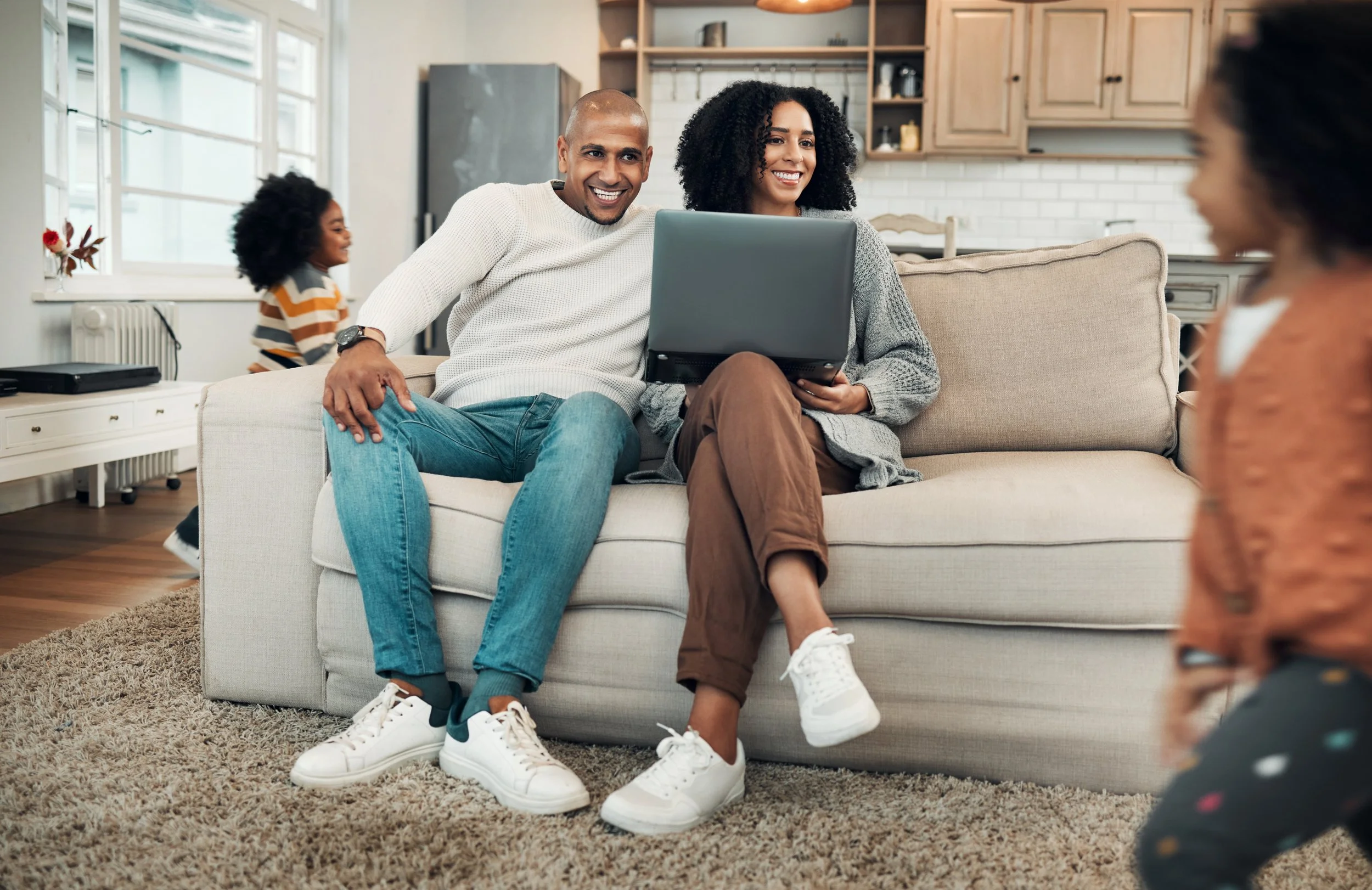 A smiling couple sitting on a sofa with a laptop, engaging with a young girl in a living room with large windows, a white brick wall, and wooden cabinets.