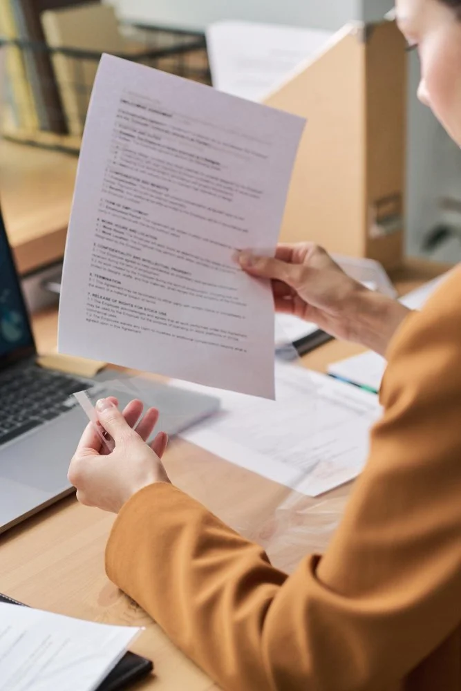 Person in an orange blazer holding a printed document in an office setting with a laptop and other papers on the desk.