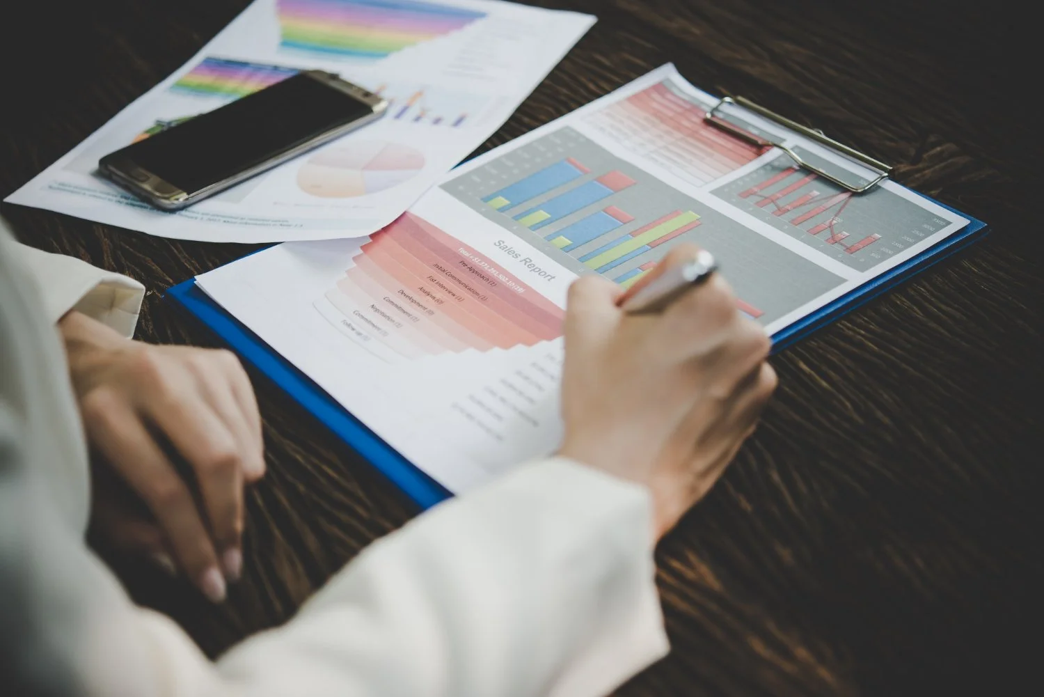 Person analyzing printed sales and performance reports with bar charts and graphs on a clipboard, on a dark wooden desk.