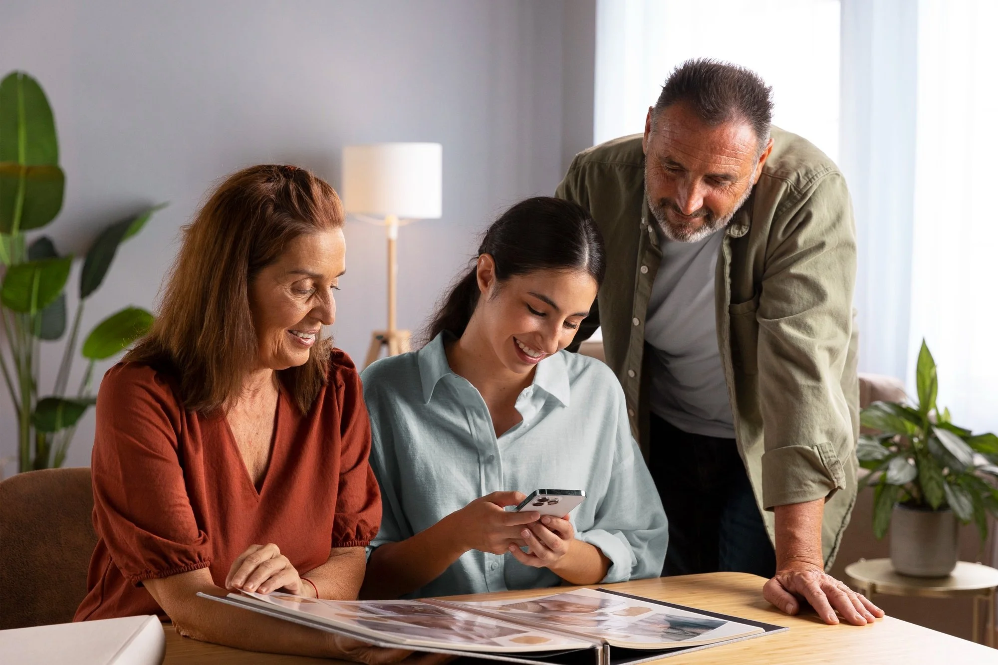 Three women and one man looking at photographs and a phone together in a well-lit living room with plants.