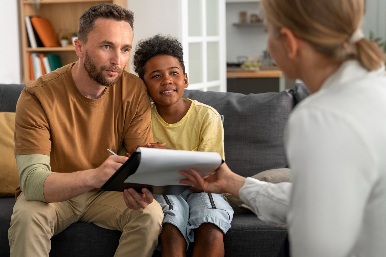 A man and a boy sitting on a sofa, talking to a woman in a white coat during a consultation.