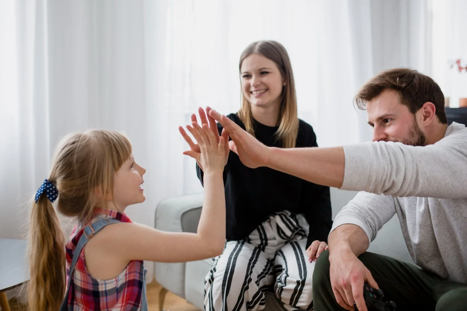A family of three having a high five in a cozy living room with white curtains