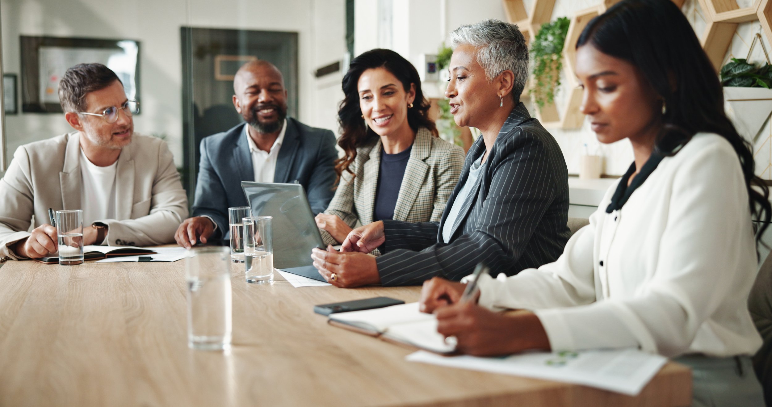 A diverse group of six professionals sitting around a conference table in a modern office, engaged in a discussion. One woman is holding a tablet, and there are glasses of water, notebooks, and a smartphone on the table.