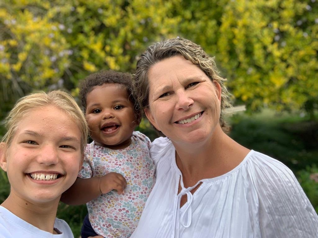 A smiling woman with two young girls outdoors in front of green trees.