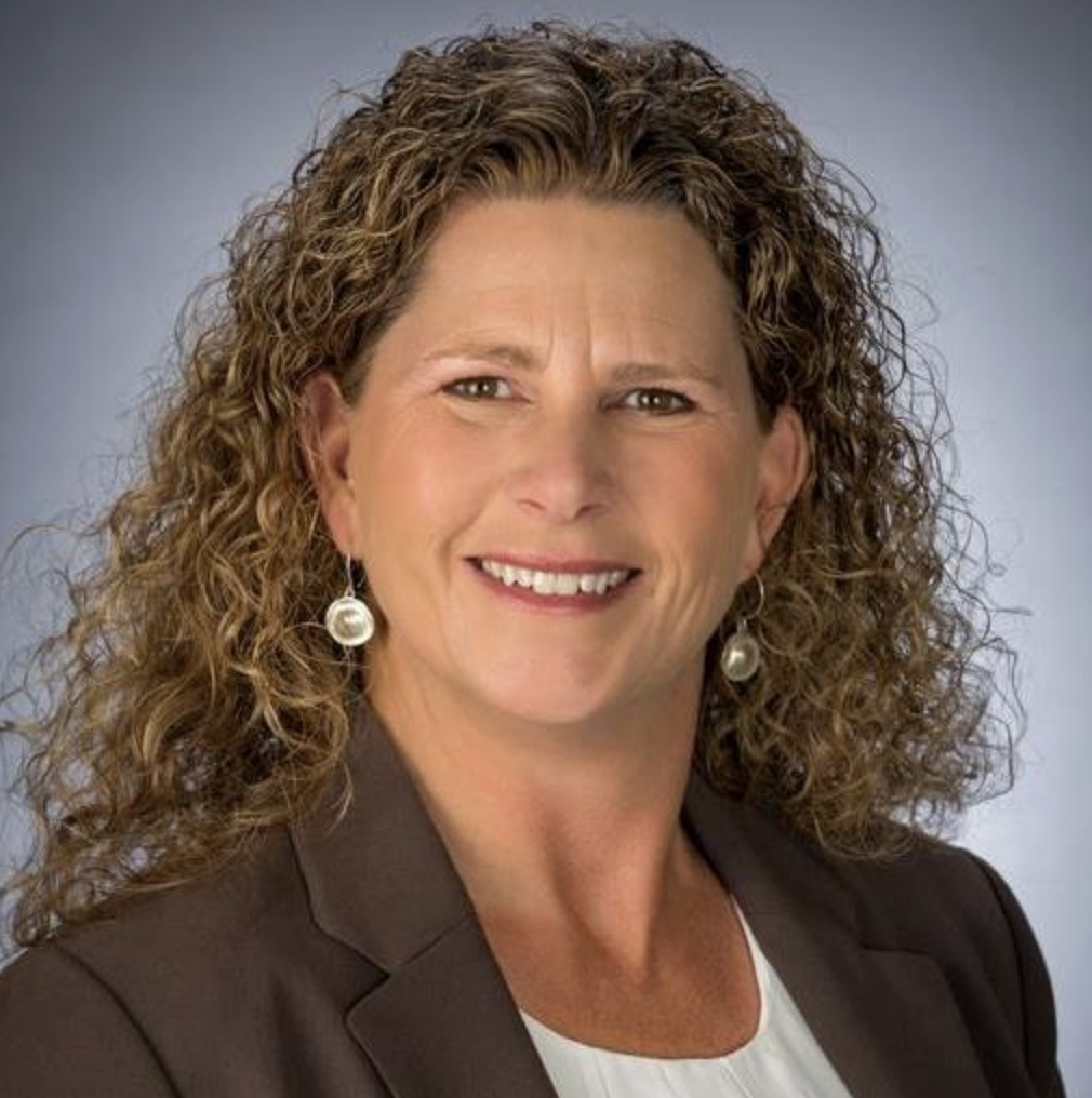 A professional woman with curly brown hair, wearing a dark blazer, white top, and pearl earrings, smiling against a gray background.