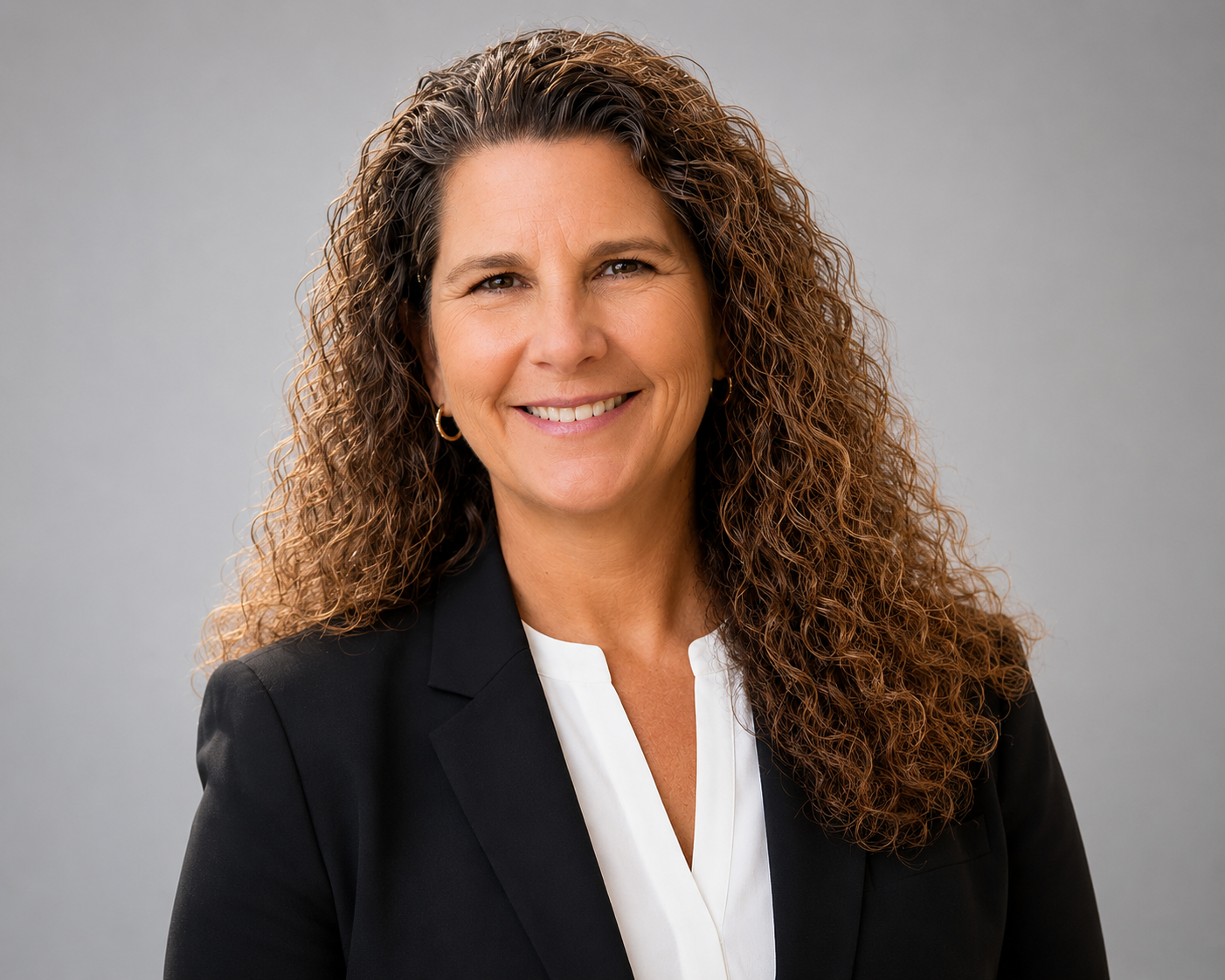 A professional woman with curly brown hair, smiling, wearing a black blazer and white top, standing against a neutral gray background.