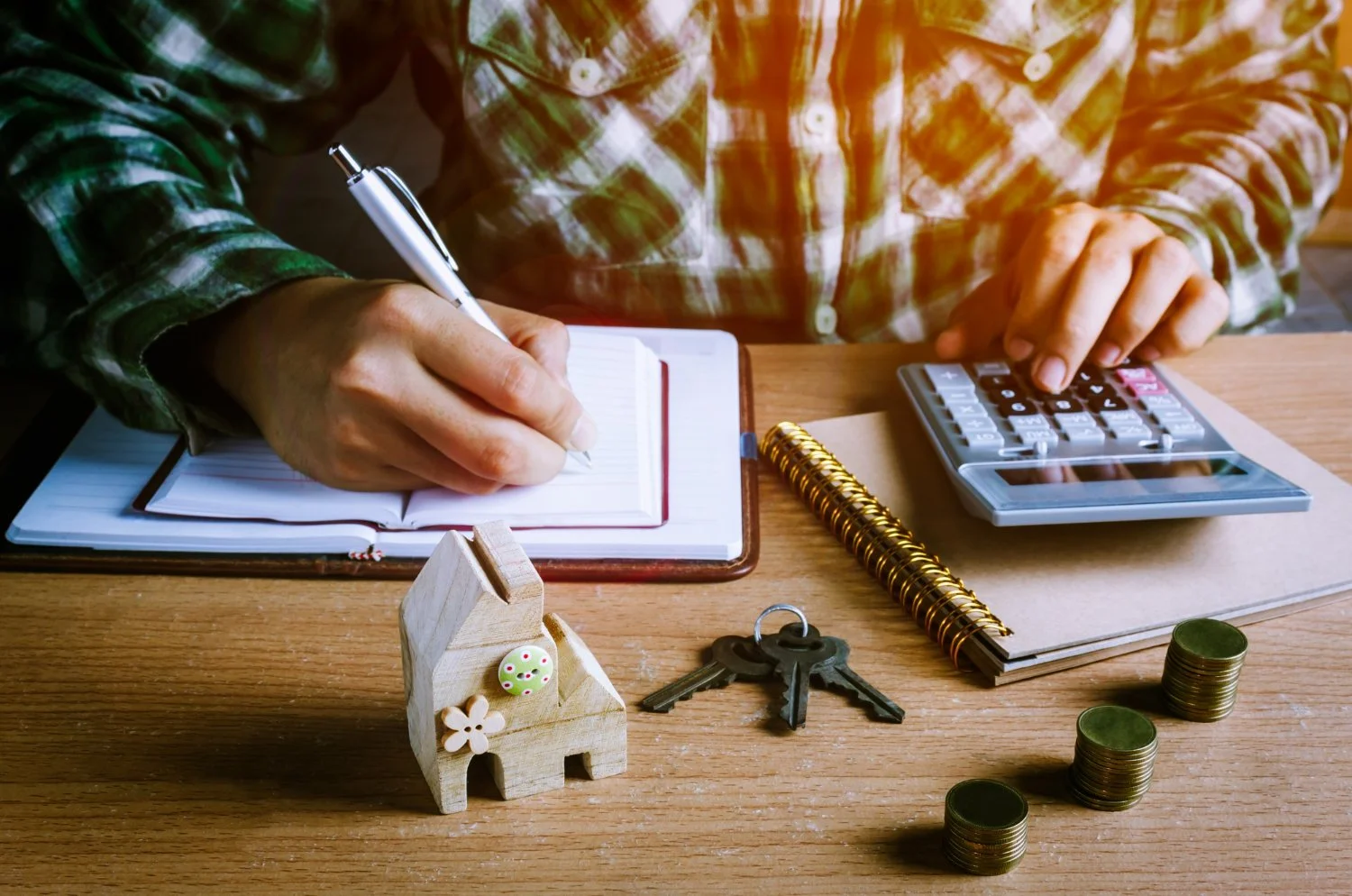 Person in plaid shirt writing in notebook with a pen, using calculator, with keys, small house figurine, and stacks of coins on wooden table.