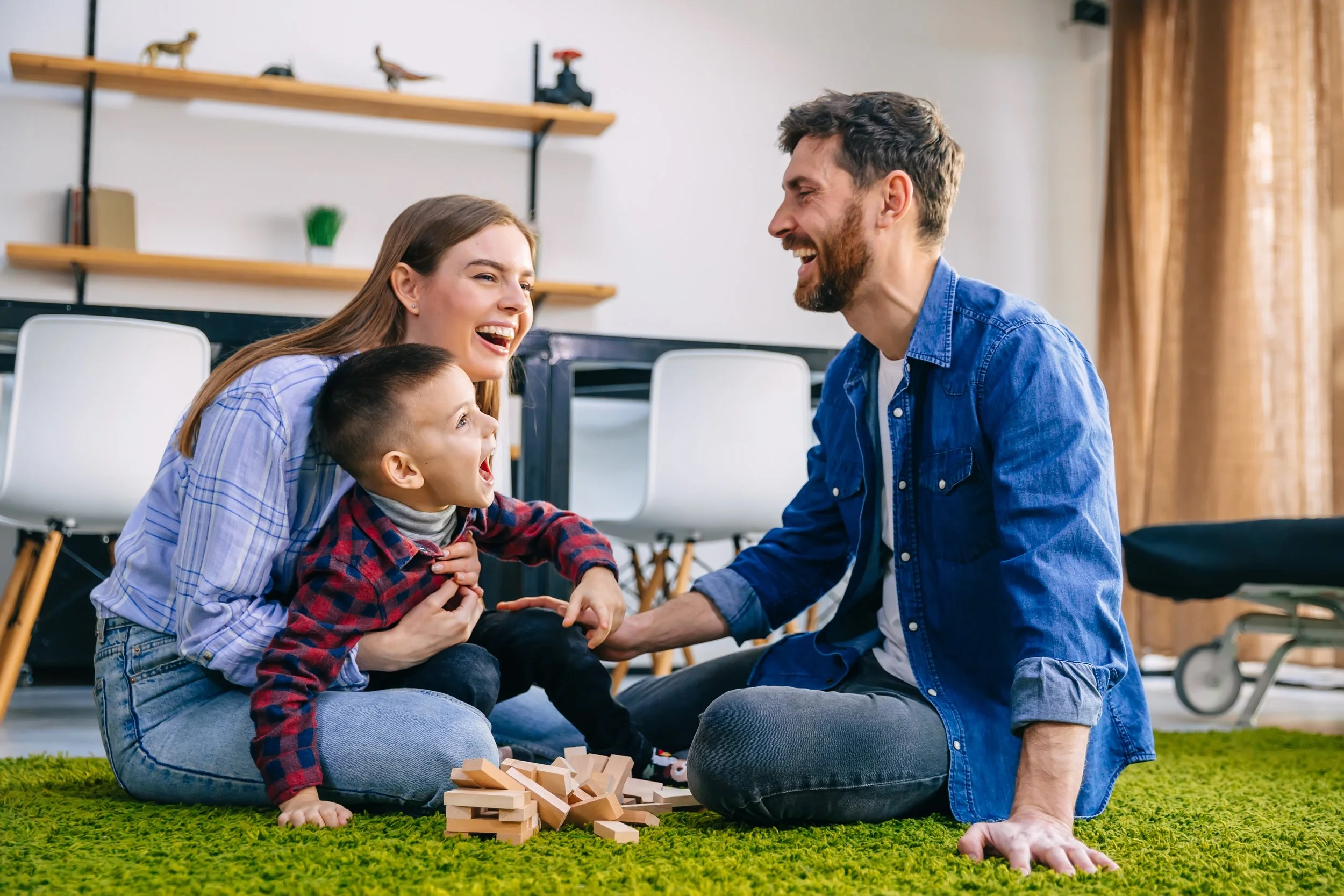 Family playing Jenga on a green carpet indoors, smiling and laughing.