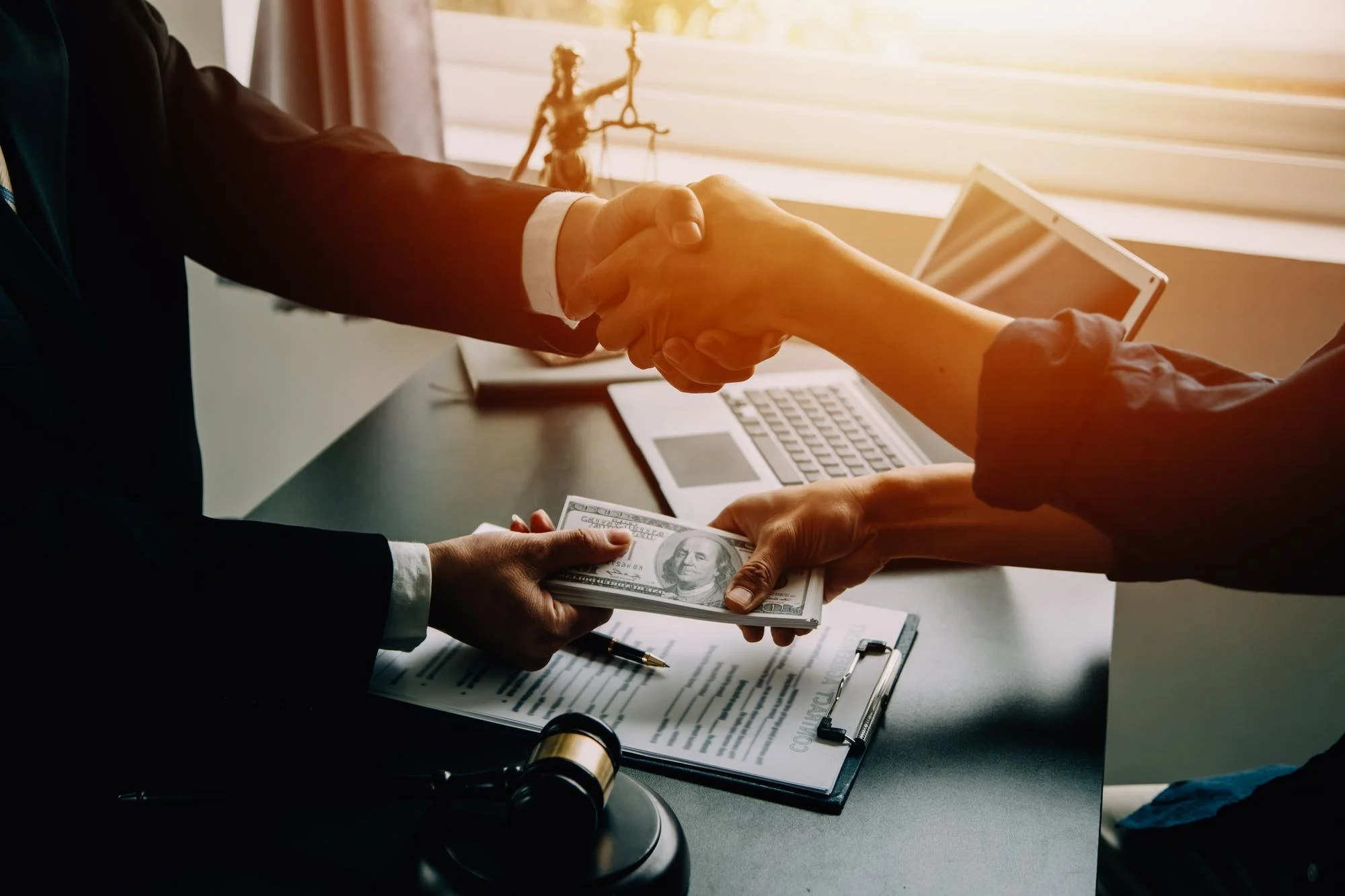 Two people shaking hands over a stack of hundred-dollar bills during a business transaction. A laptop, clipboard with paperwork, and pen are on the table, with a lady justice statue in the background.