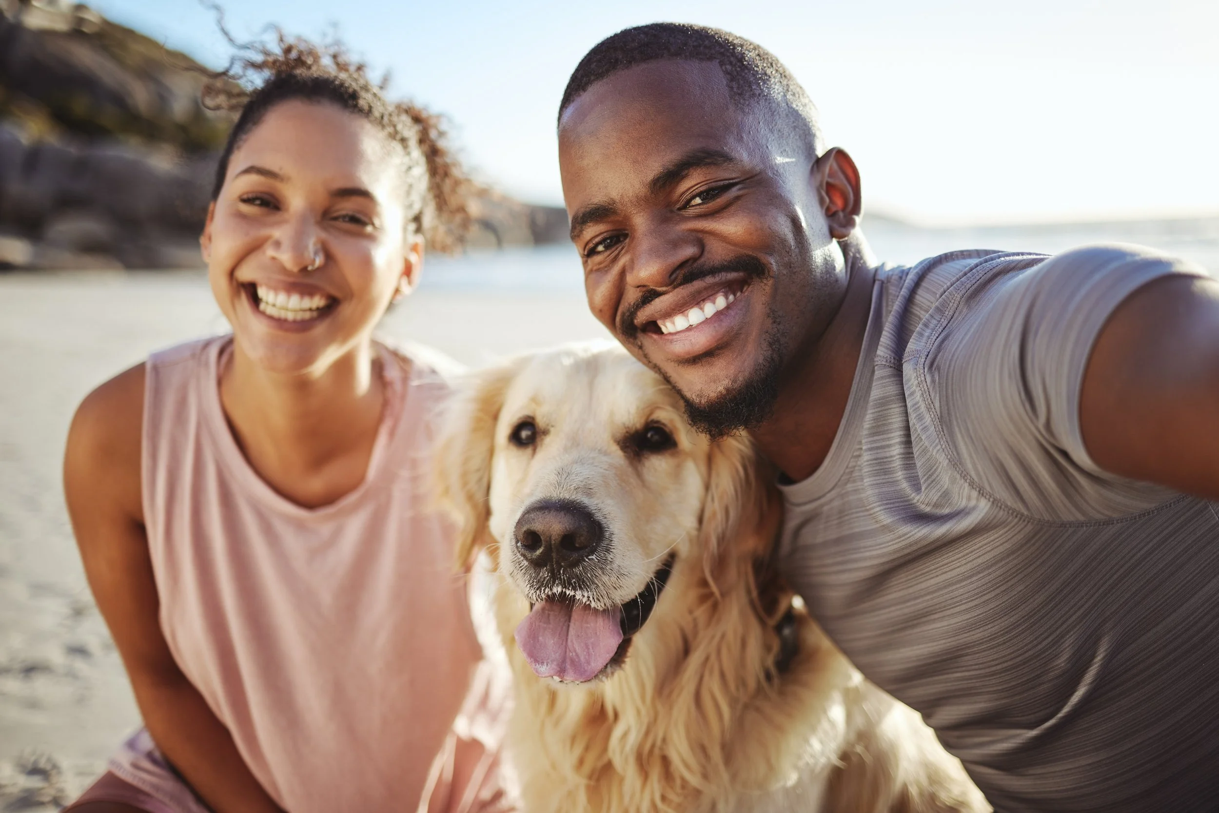A smiling young woman and man taking a selfie at the beach with a golden retriever dog, ocean in the background.