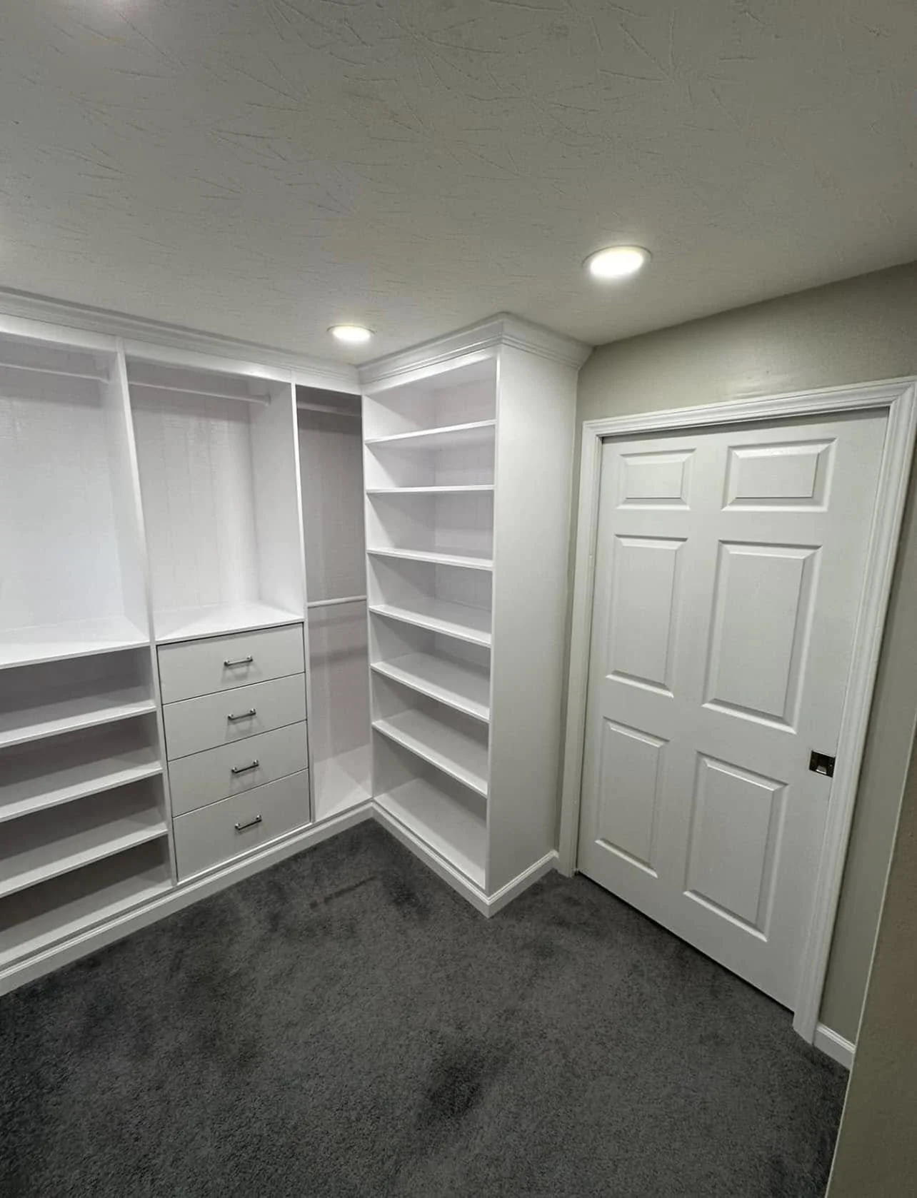 Empty white walk-in closet with shelves, drawers, and a closed door, with the ceiling lights turned on.