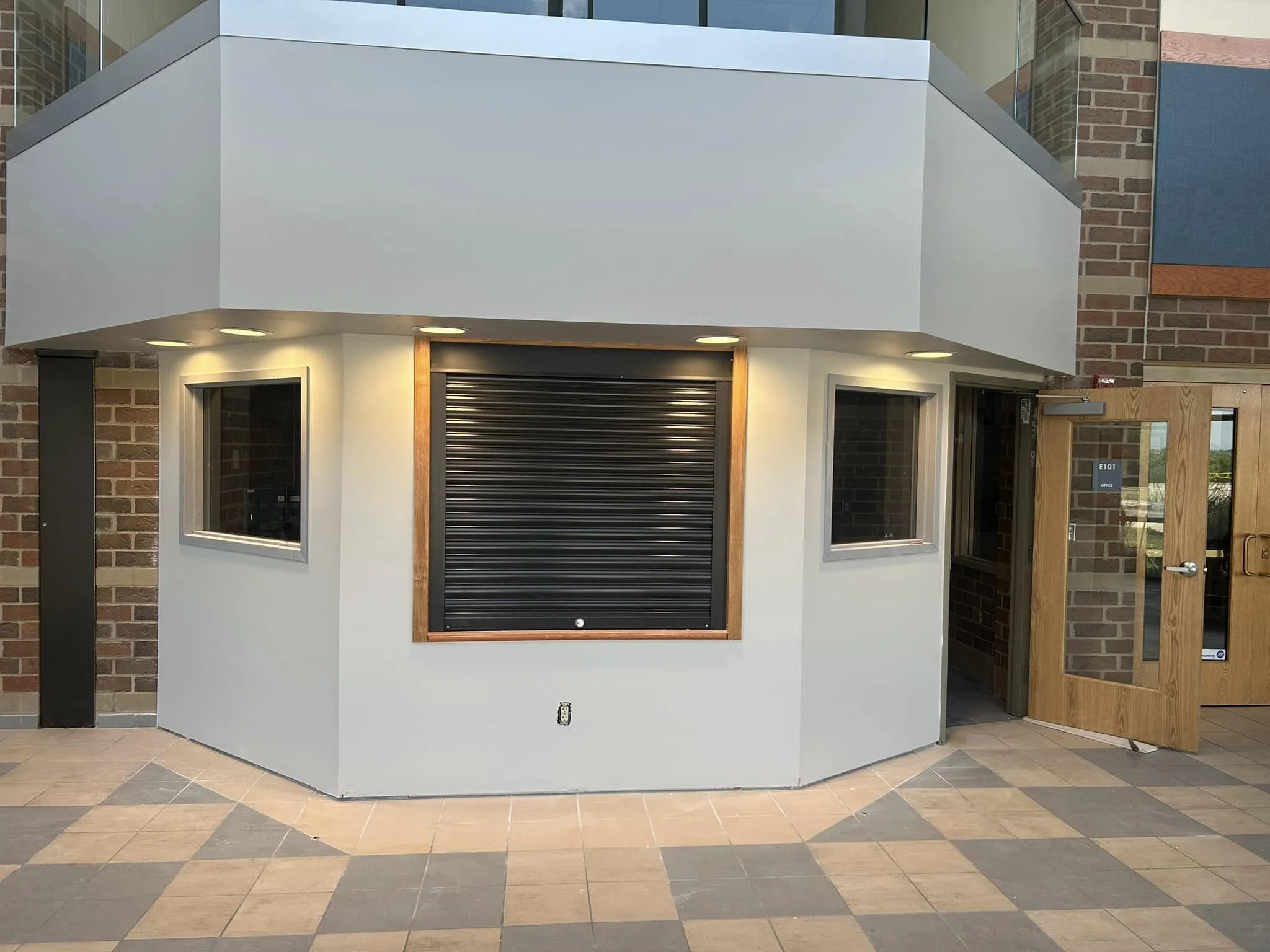 A small building with a modern design, featuring a white exterior, a black roll-up door, and two small windows. The top part of the structure extends outward with recessed lights underneath. The surrounding wall is made of bricks, and a wooden door with a glass window and a metal handle is to the right of the building. The floor is tiled with a pattern of dark and light beige tiles.