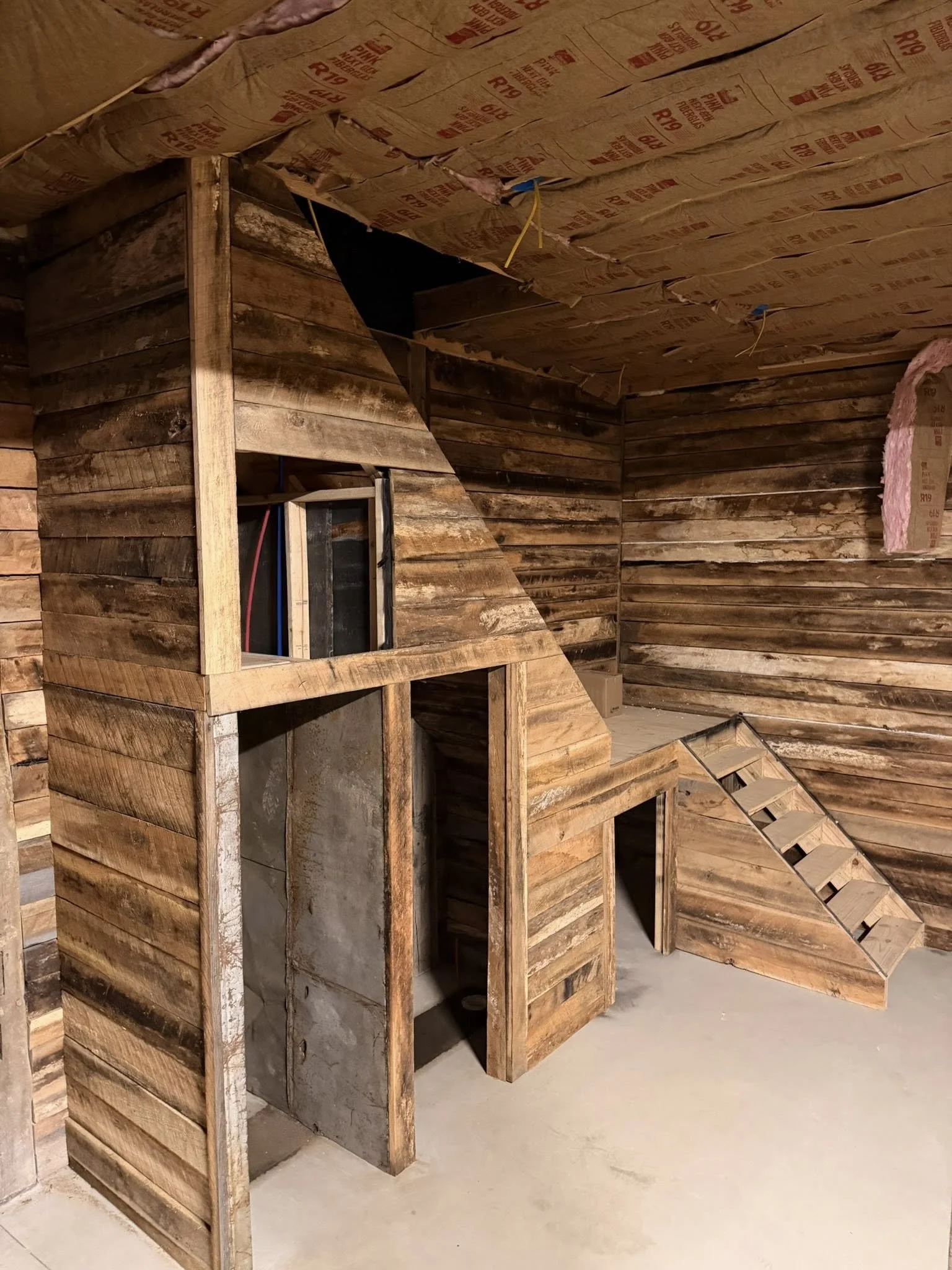 Interior of a building under construction with wooden framing and insulation, including a staircase and a small doorway.