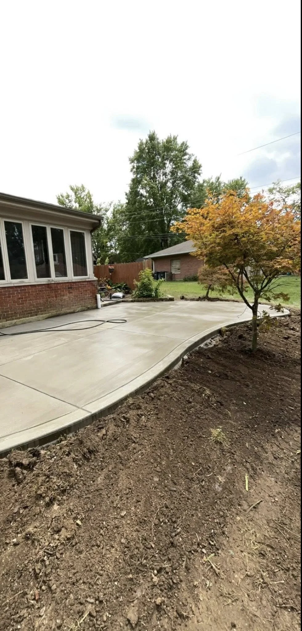 Photo of a backyard with a fresh concrete patio, a small tree with orange leaves, and a house with a sunroom. The yard is partially landscaped with some dirt and plant beds.
