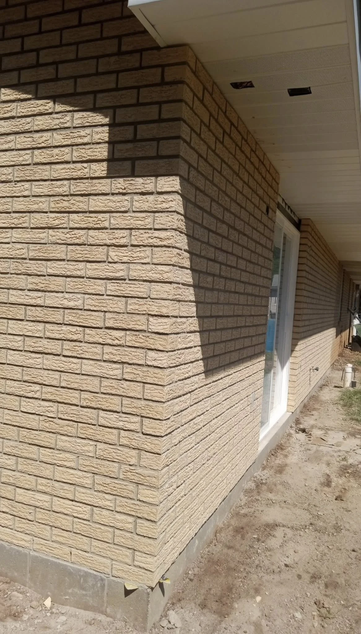 The exterior of a house with beige brick walls and a white sliding glass door, showing part of a backyard with dirt ground.