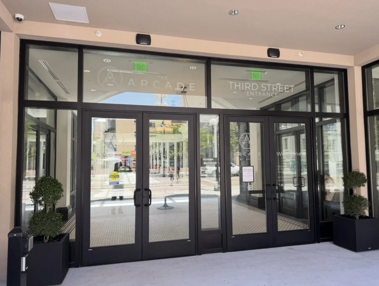 Glass entrance doors with the signs 'The Arcade' and 'Third Street Entrance' above them, surrounded by two potted plants inside and a city street outside.