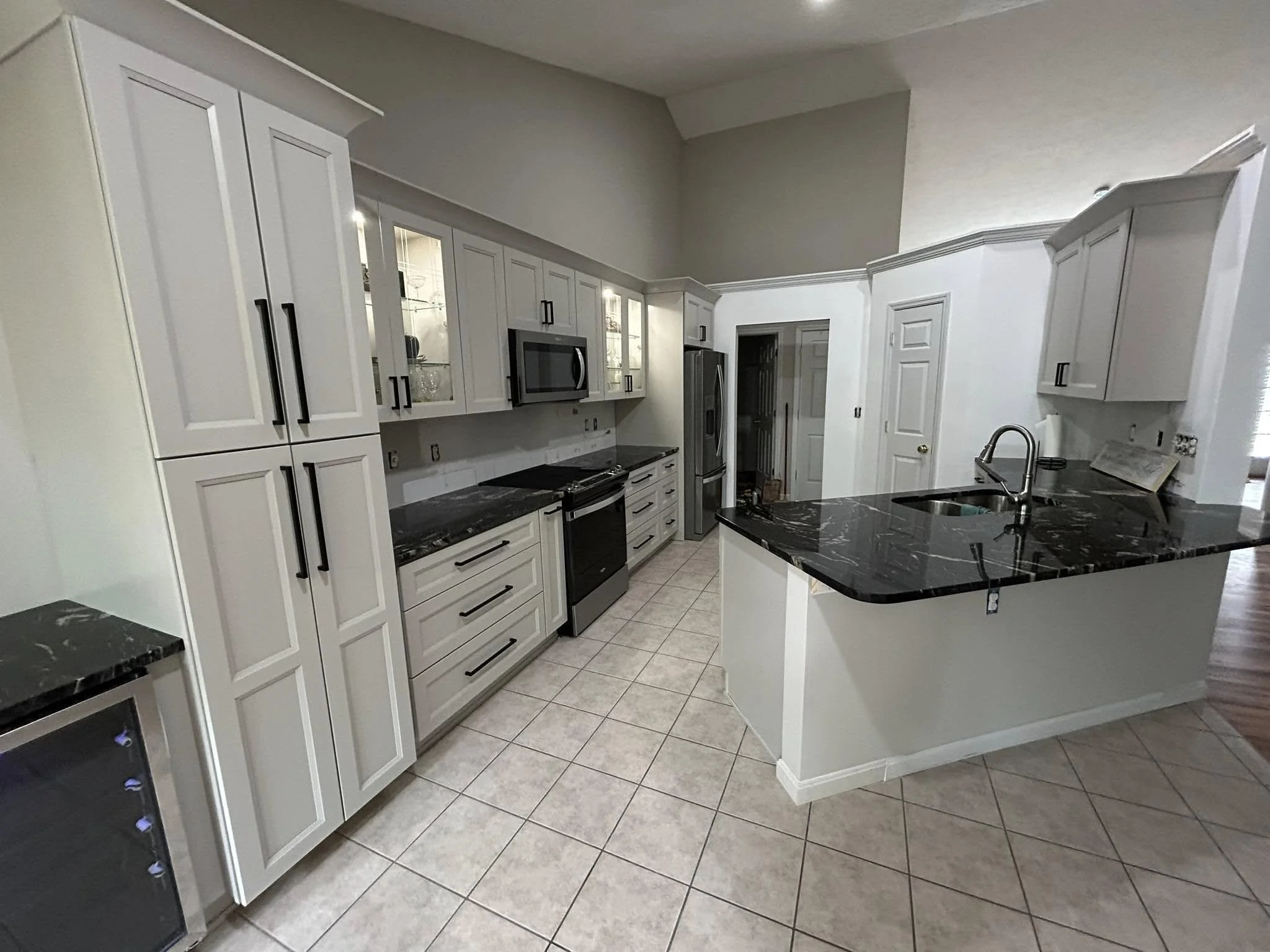 A kitchen with white cabinets, black countertops, a stainless steel microwave, oven, and refrigerator, and a tiled floor.