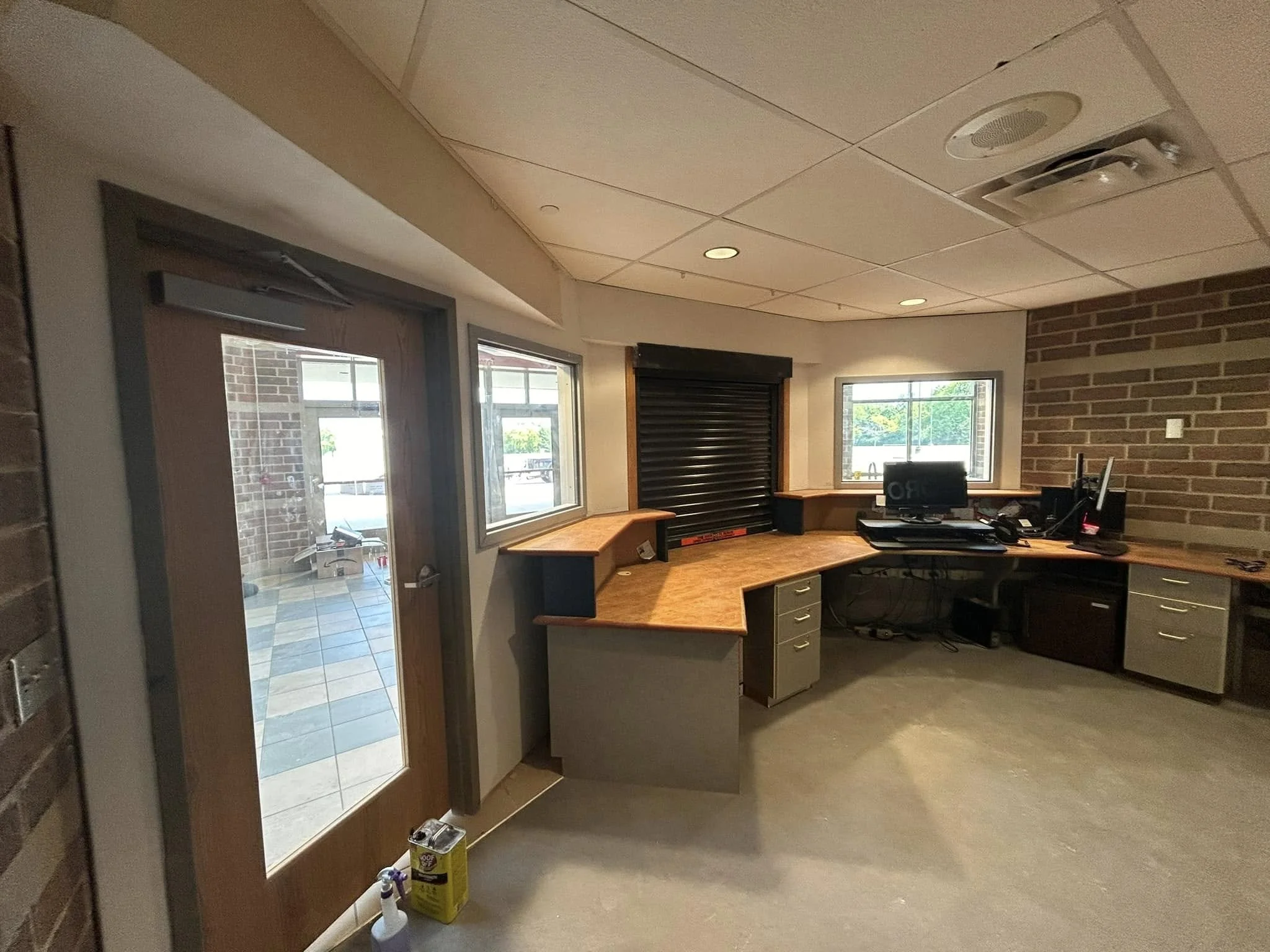 Interior of a room with a corner of a kitchen or office area, including a curved wooden counter, computer monitor, and office supplies. Outside the door, there is a tiled area with a brick exterior wall visible through the glass door and windows.