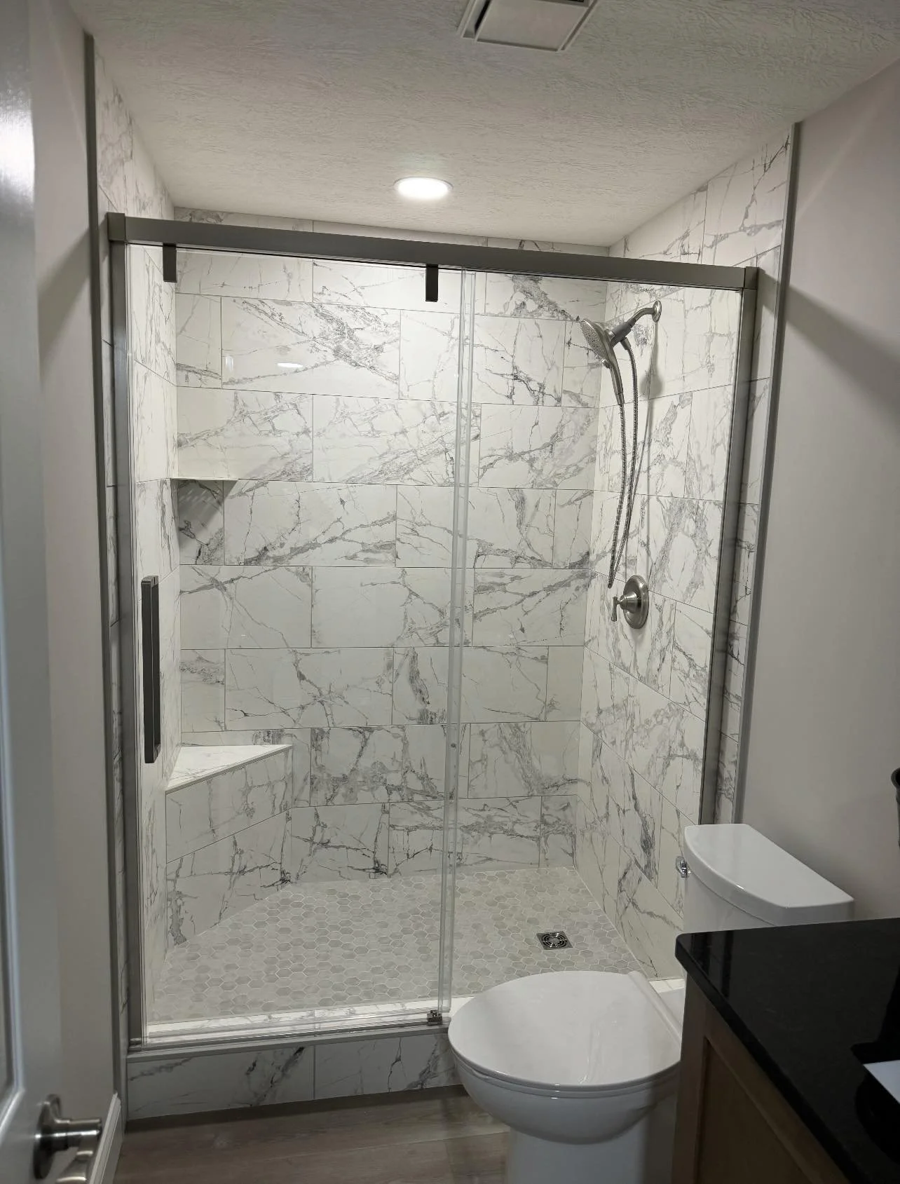 Modern bathroom with a marble shower, glass door, and a toilet. The shower features a built-in bench and marbled tiles on the walls and floor.