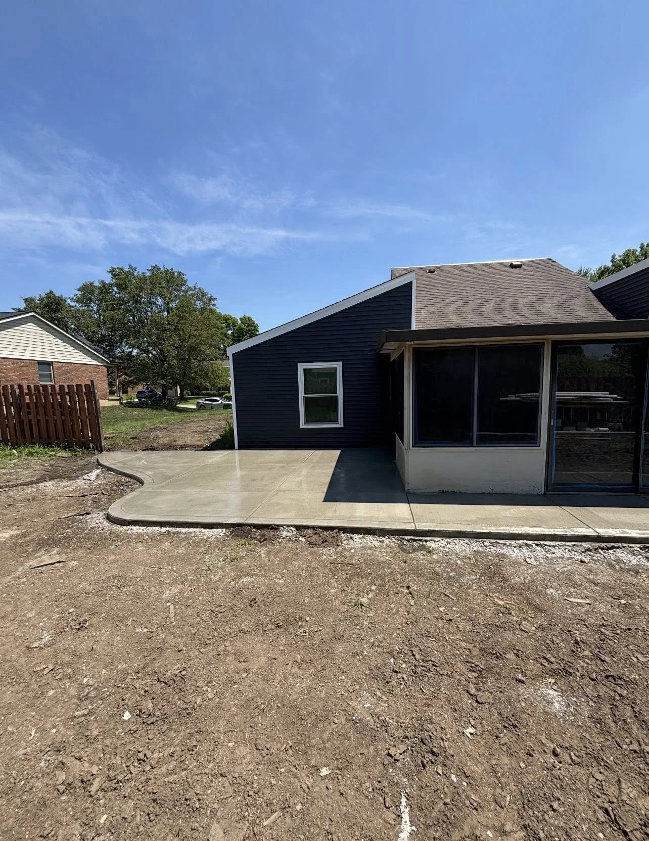 Newly poured concrete patio with curved edge in backyard of a house, with some dirt and construction area in the foreground, a dark blue house with large windows and a screened porch attachment, a neighboring brick house, trees, and a clear blue sky.